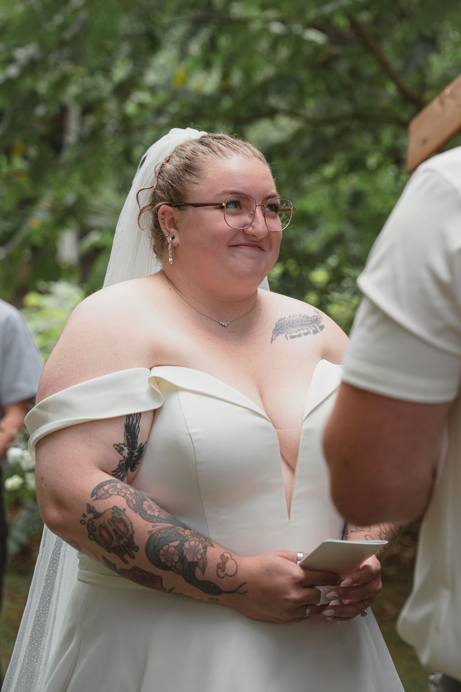 Bride in white off-shoulder gown with tattoos and veil smiles during outdoor wedding ceremony.