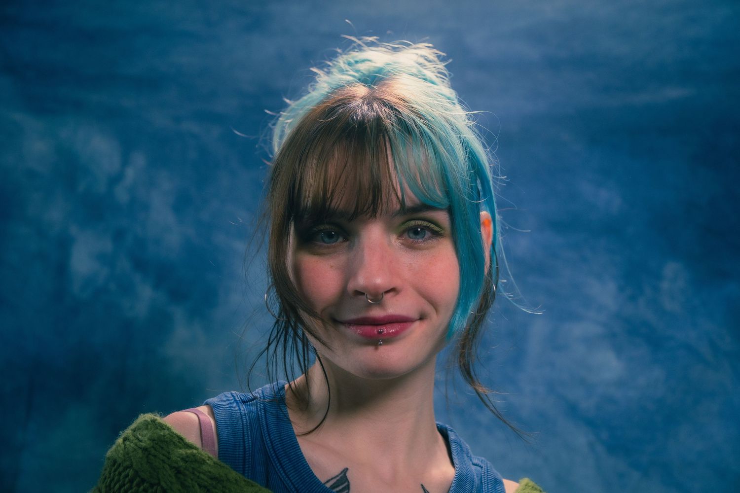 Young woman with teal highlighted bangs and updo smiling softly in atmospheric blue studio portrait.