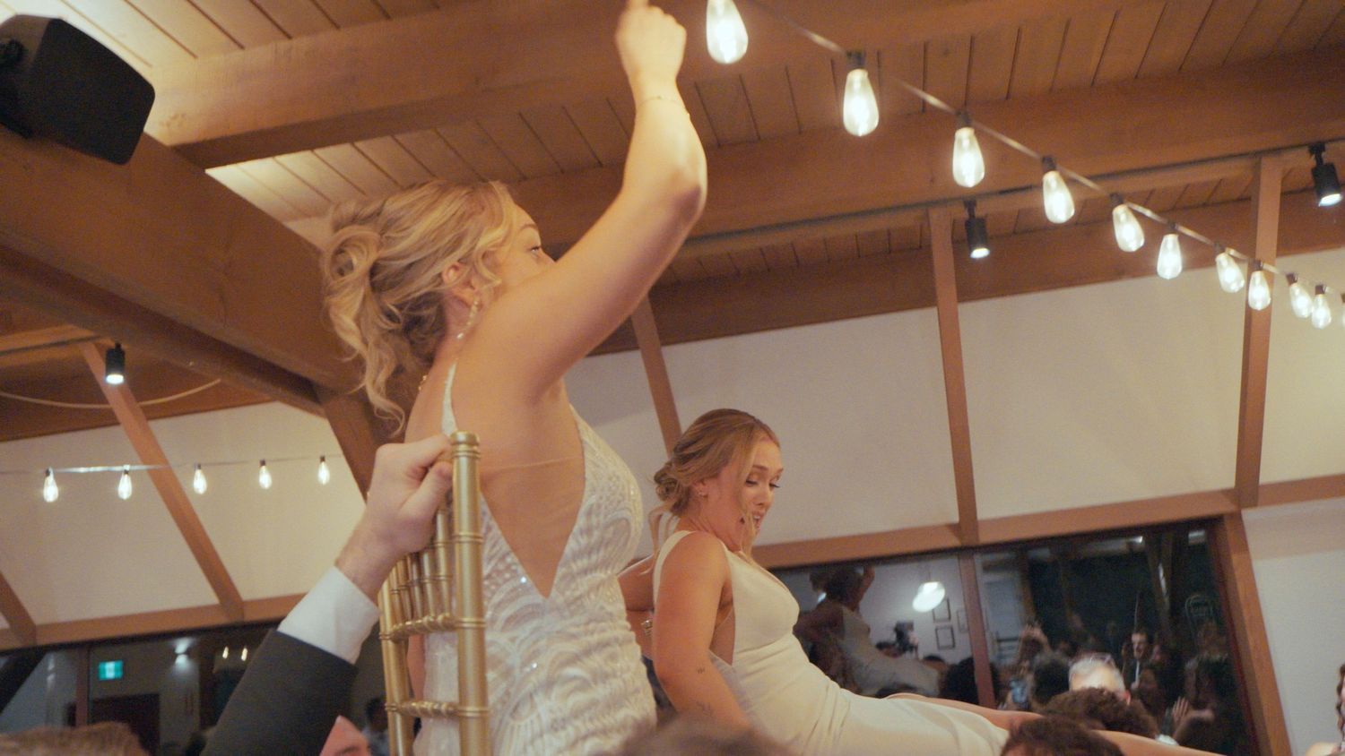 Bride being lifted in chair during joyful wedding reception dance celebration in decorated venue with string lights.