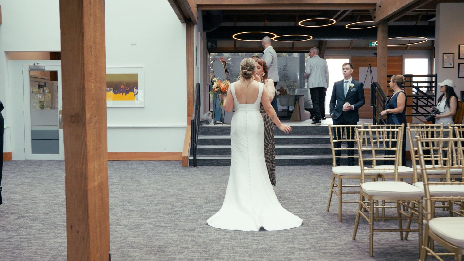 Bride in white wedding gown stands at venue aisle with gold chairs before ceremony.