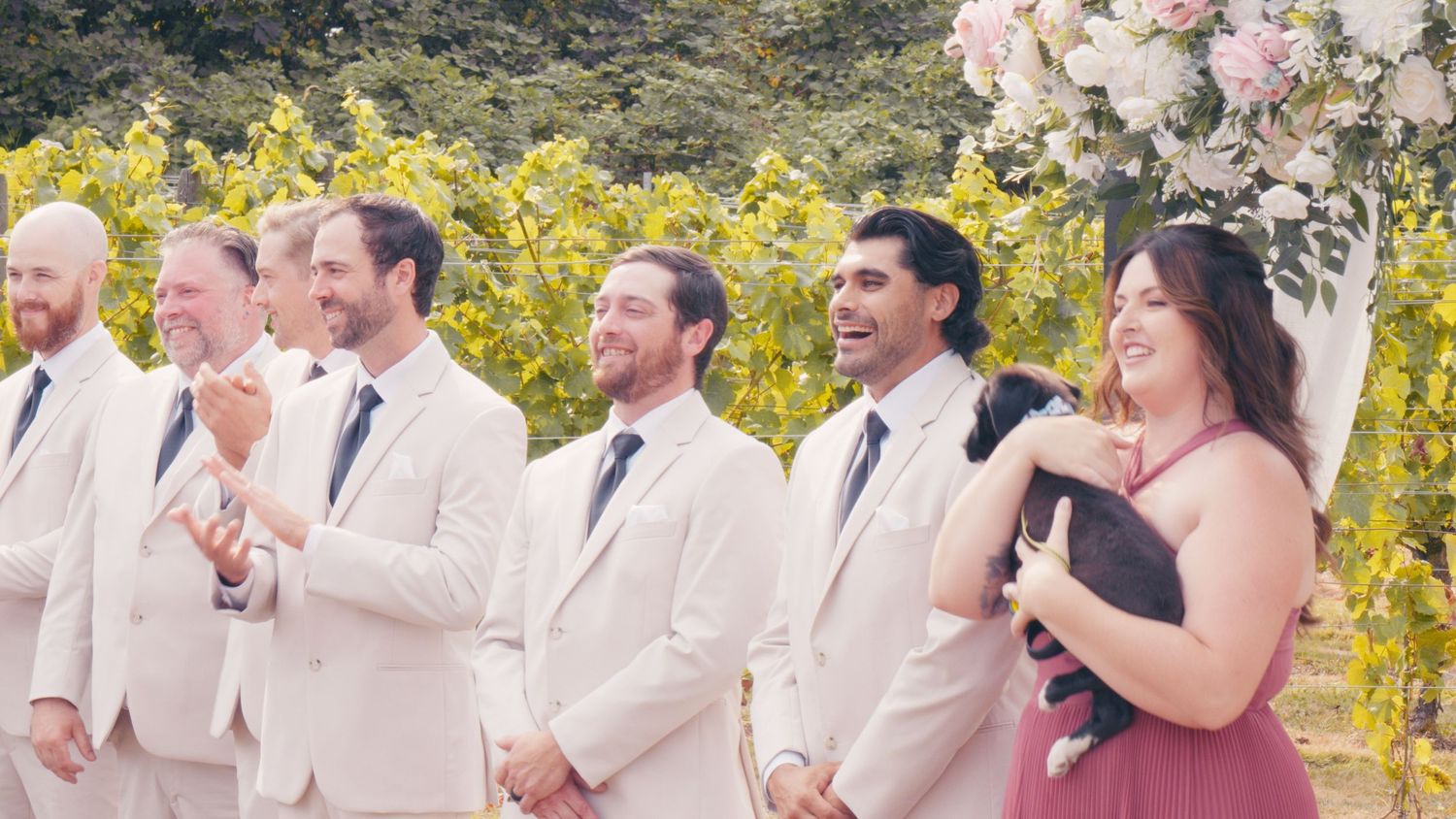 Wedding party in cream suits and pink bridesmaid dress laugh and celebrate at outdoor vineyard ceremony.