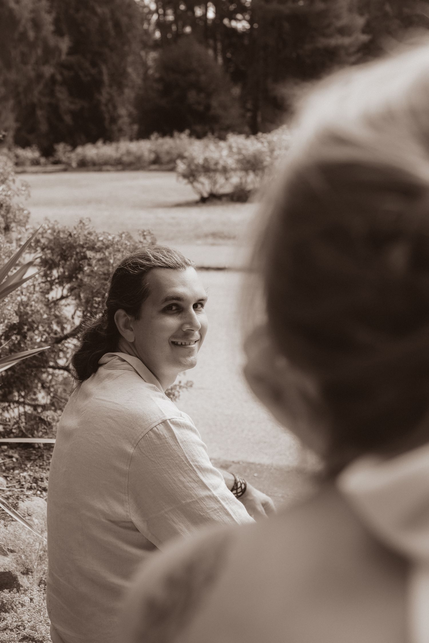 Smiling groom looks back at bride in a candid sepia-toned outdoor wedding portrait among garden greenery.