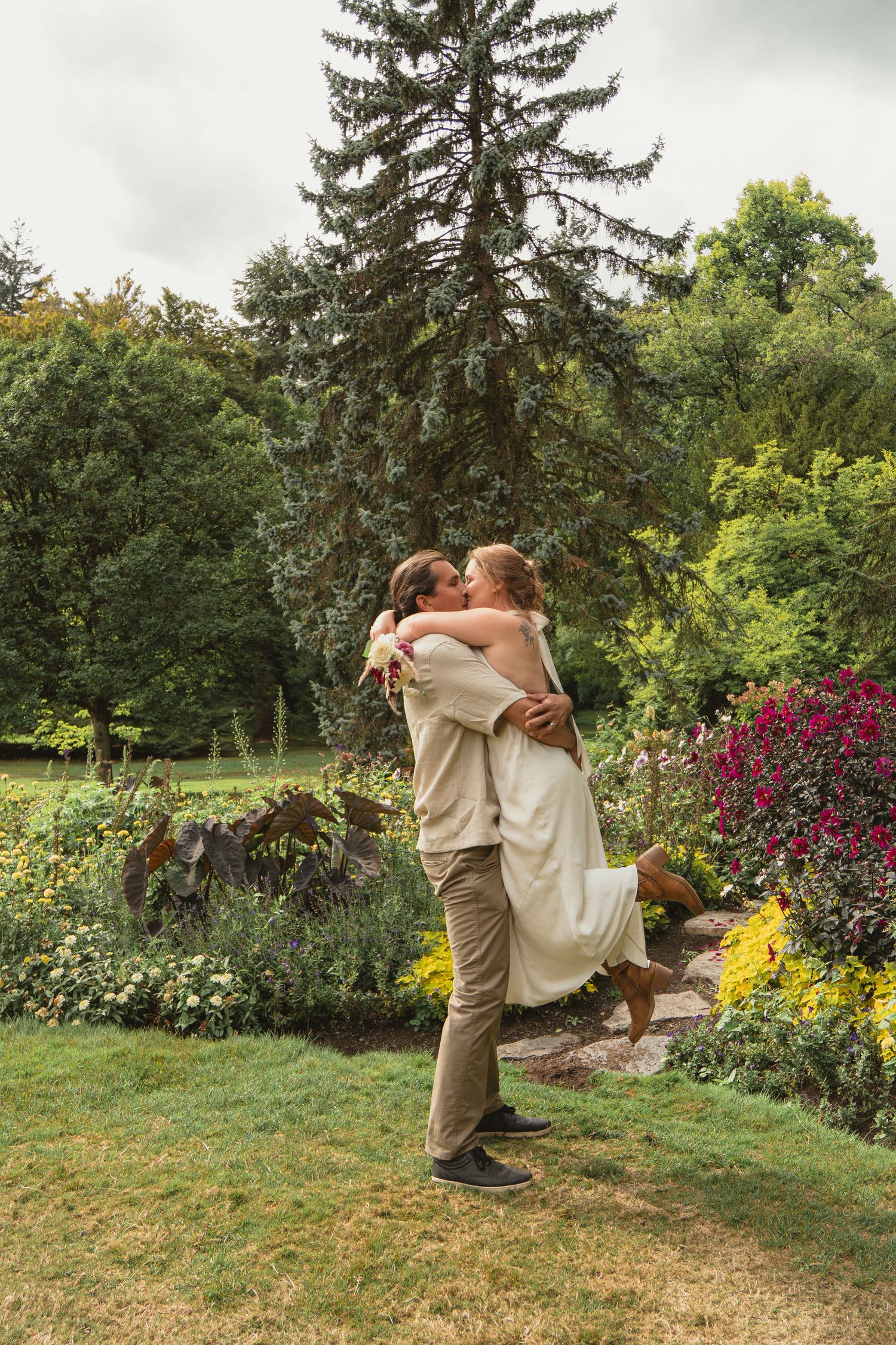 Groom lifts and embraces bride in white dress surrounded by vibrant flowers in a lush garden.