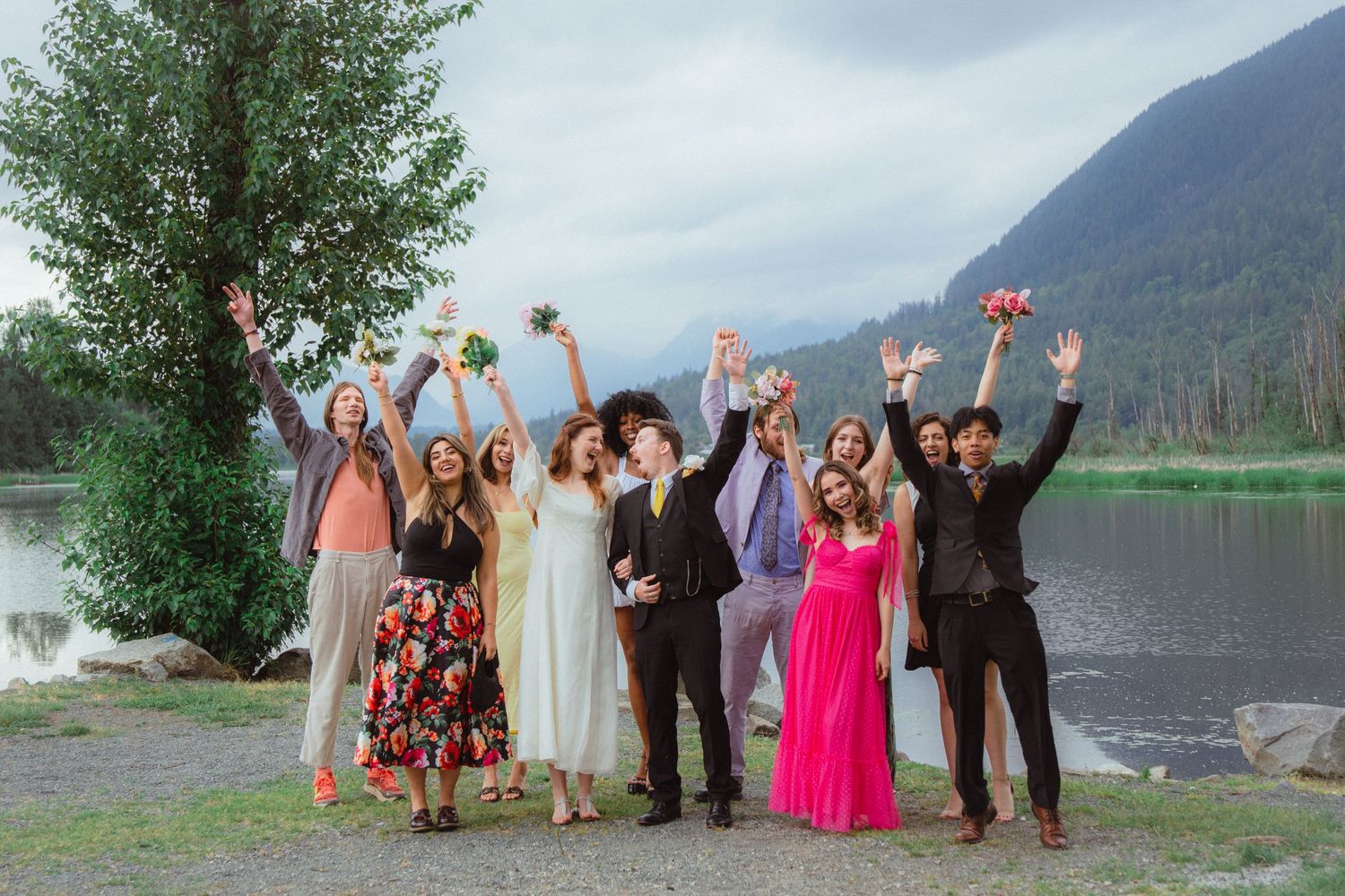 Joyful wedding party celebrating with raised hands and bouquets near a scenic mountain lake.