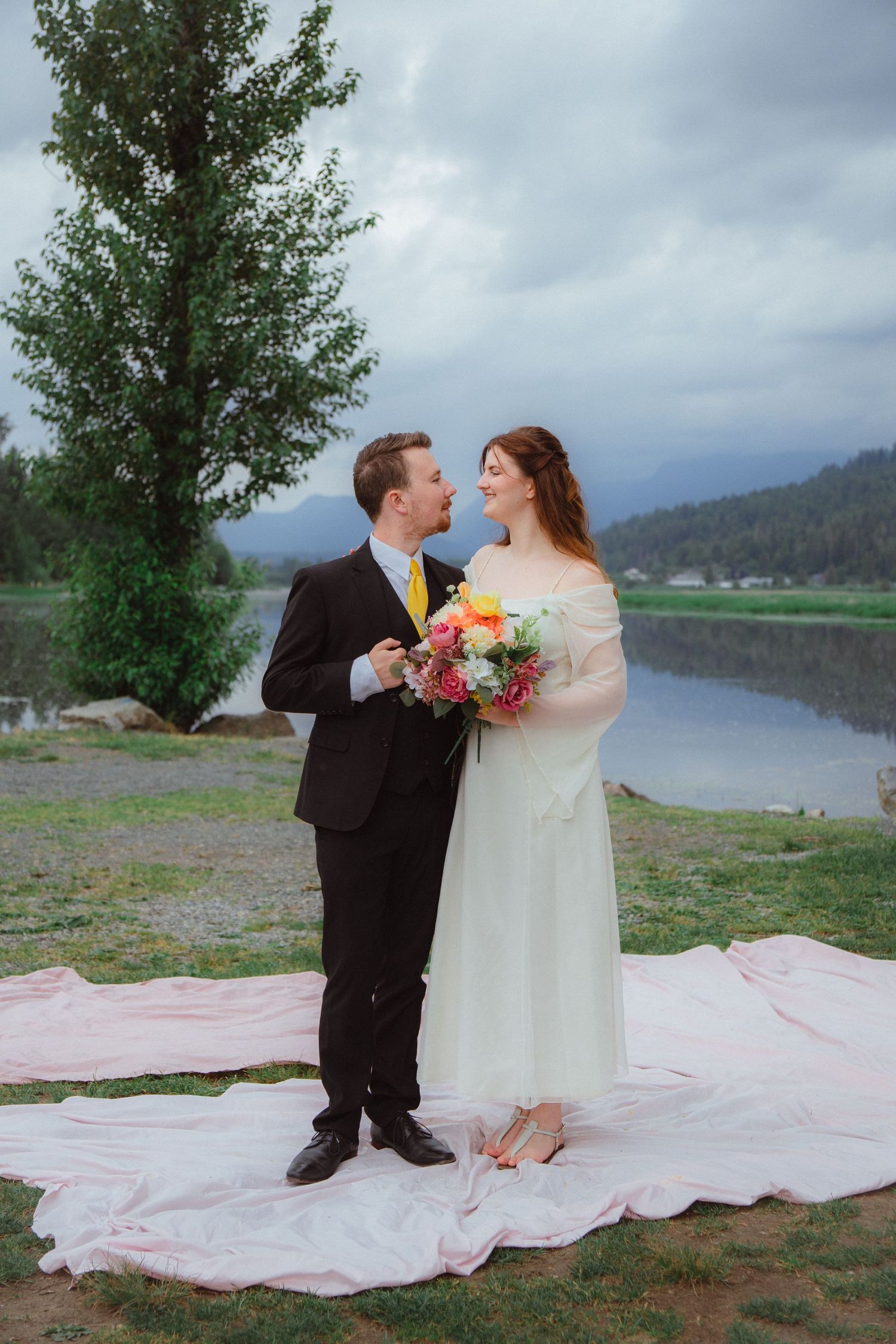 Bride and groom share a joyful moment on pink fabric by a scenic mountain lake.