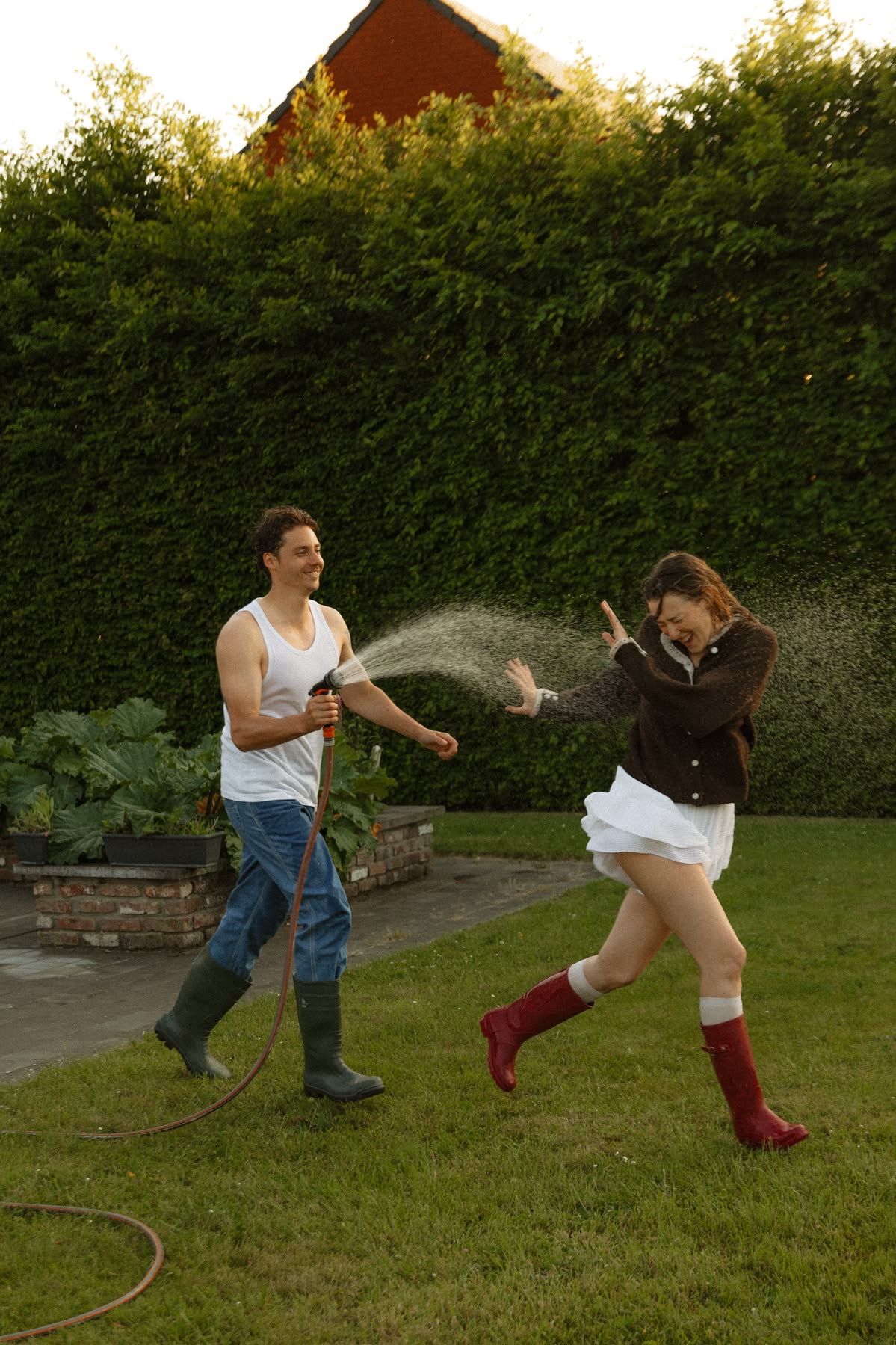 A man sprays a laughing woman with a garden hose in a backyard, both wearing rubber boots on green grass.