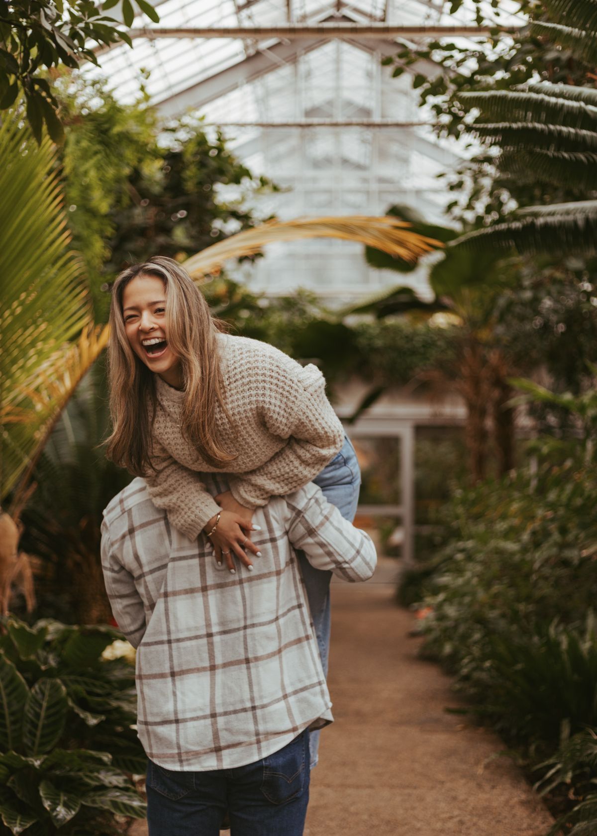 A laughing woman gets a piggyback ride through a lush greenhouse filled with tropical plants.