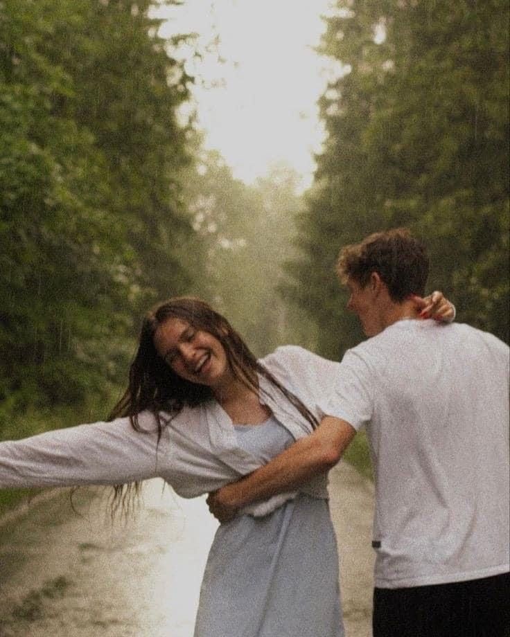 Couple dancing joyfully in the rain on a forest road, woman laughing with arms outstretched in white outfit.