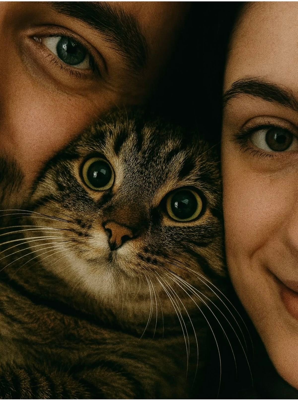 Two people press their faces close together with a tabby cat nestled between them, all three looking directly at the camera.