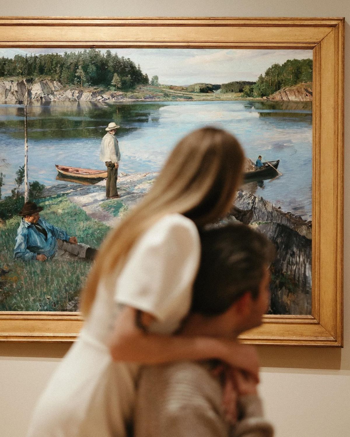 Couple embracing while admiring a large landscape painting of a serene river scene with fishermen in a museum gallery.