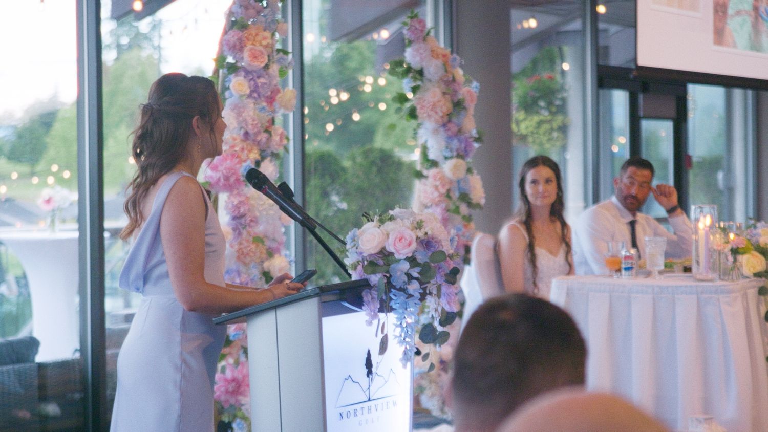 Woman gives a speech at a wedding reception podium while the bride and groom listen at the head table.