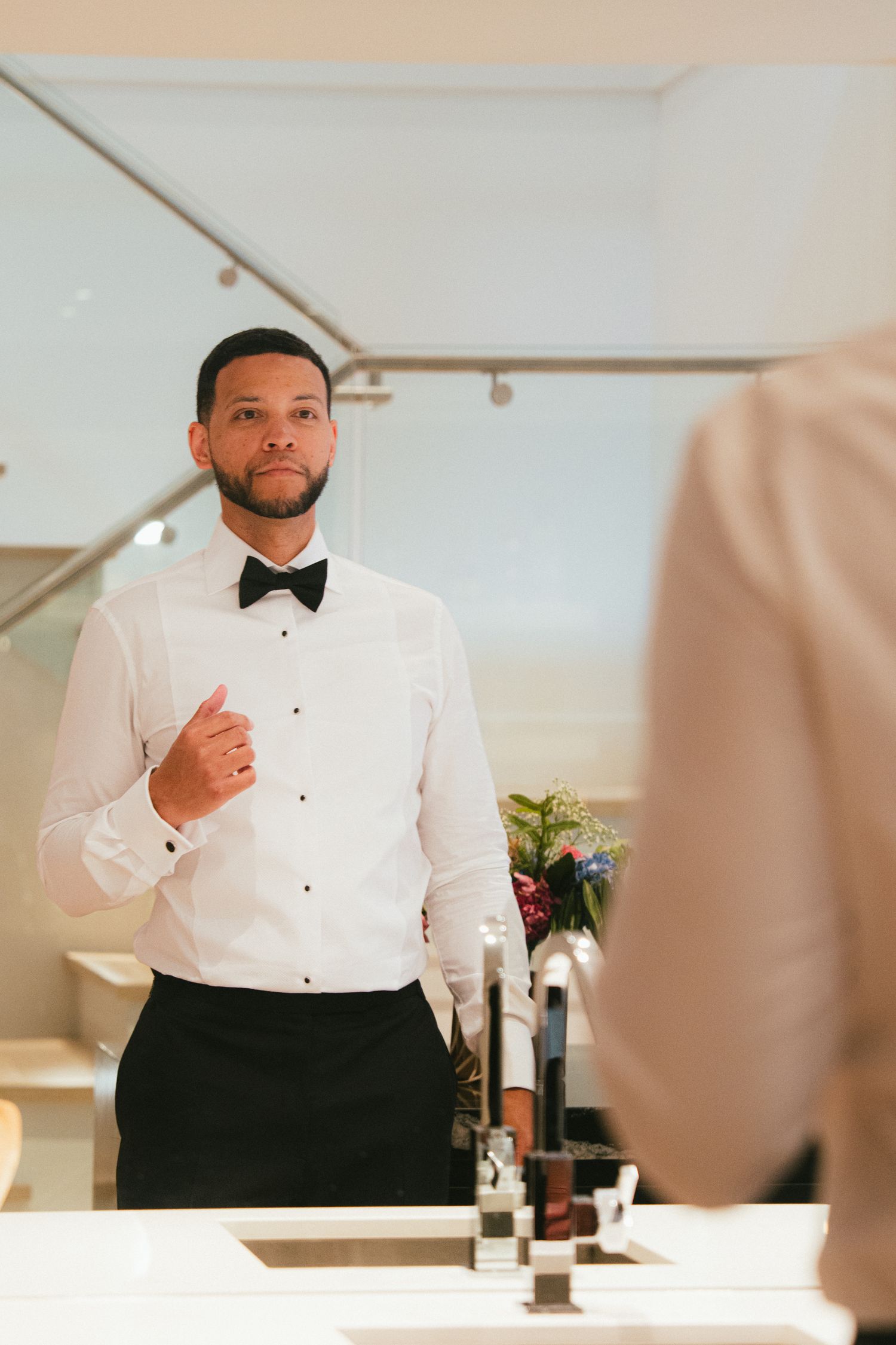 Groom in white tuxedo shirt and black bow tie checking his appearance in a bathroom mirror before wedding.