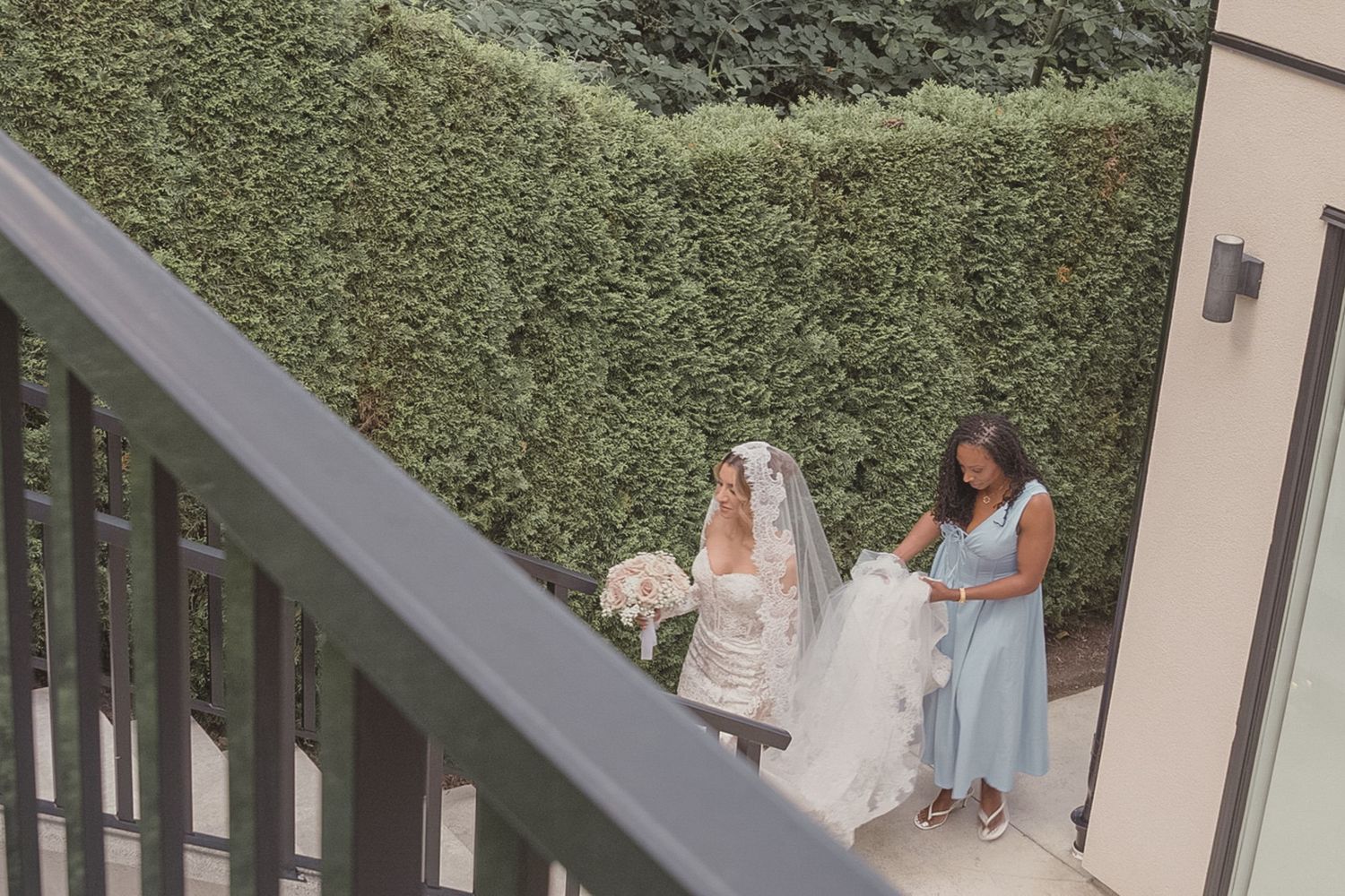 Bride in white gown and veil holding bouquet with bridesmaid in blue dress on outdoor patio near green hedge.