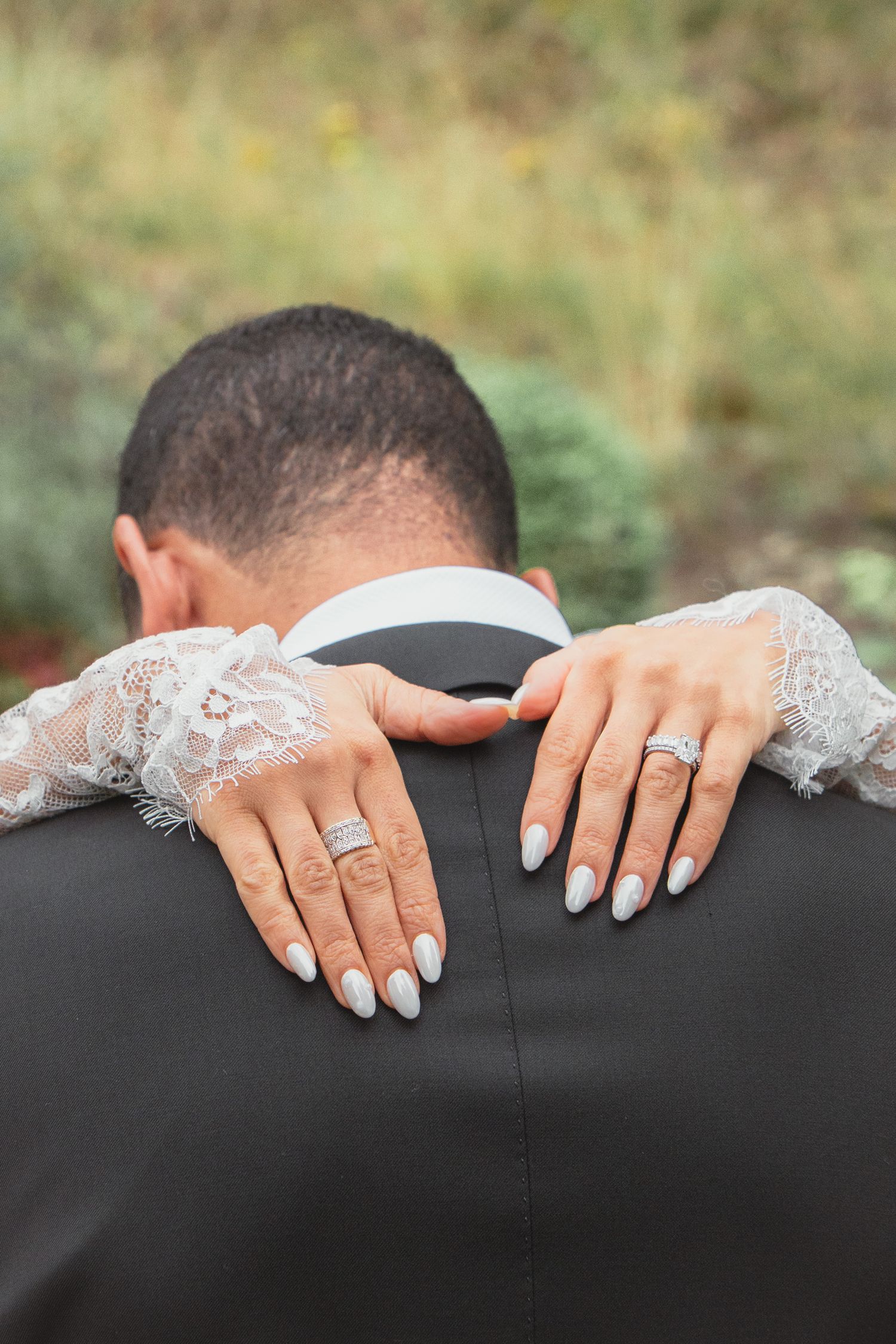 Bride with white manicure and diamond wedding ring embracing groom in black suit from behind outdoors.