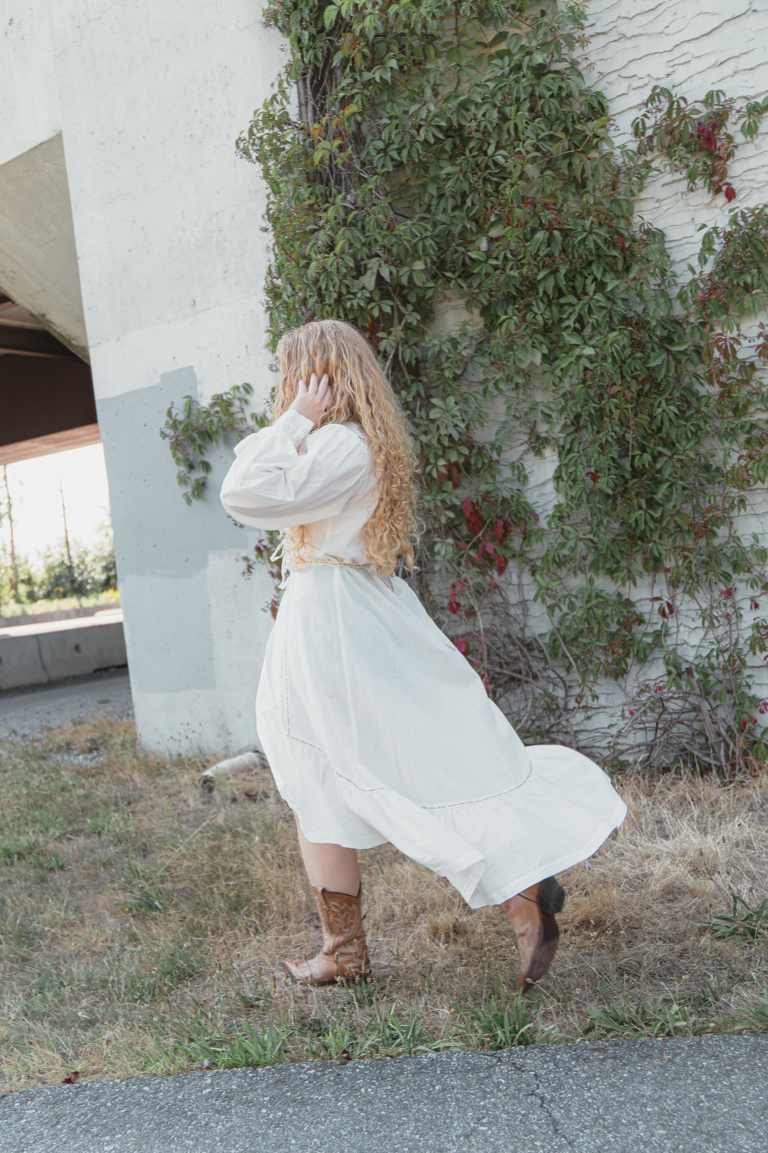 Curly blonde woman in flowing white maxi dress and brown cowboy boots stands by vine-covered wall.