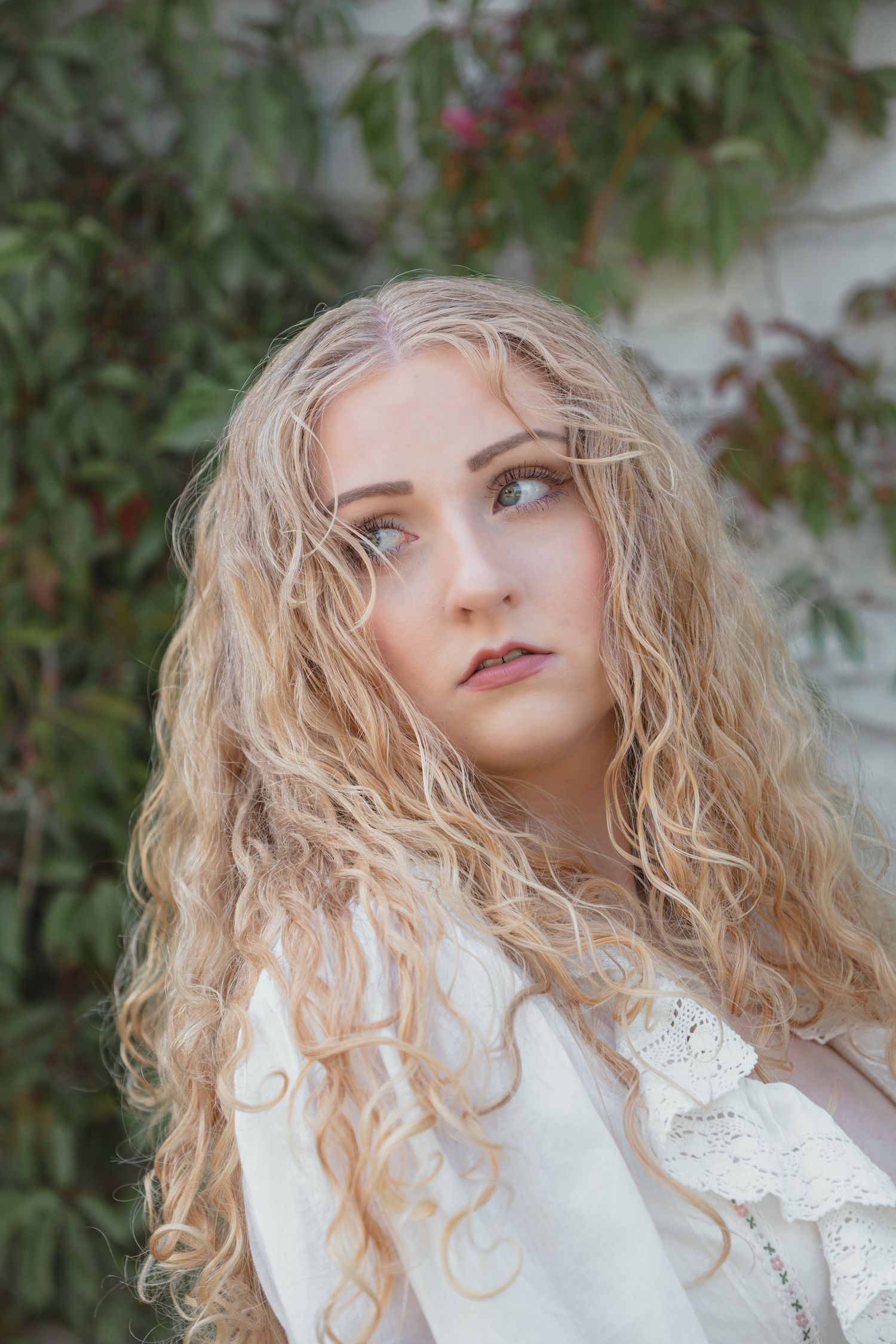 Young woman with long curly blonde hair wearing white lace top, gazing upward against green foliage backdrop.
