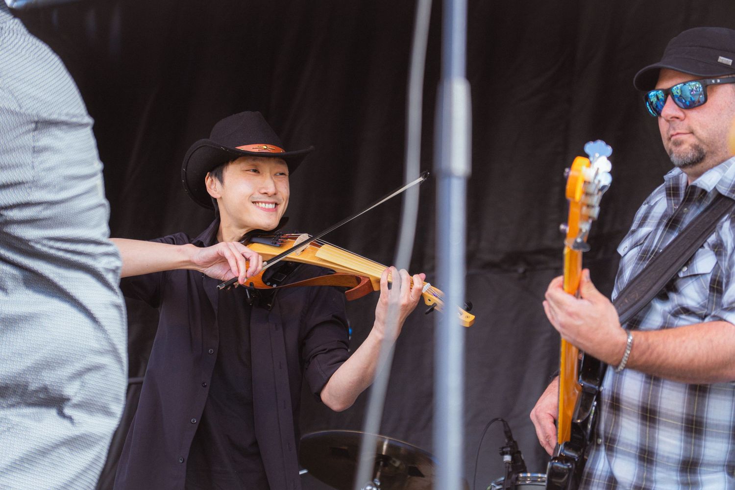 Smiling violinist in black hat plays golden electric violin alongside guitarist at outdoor concert.