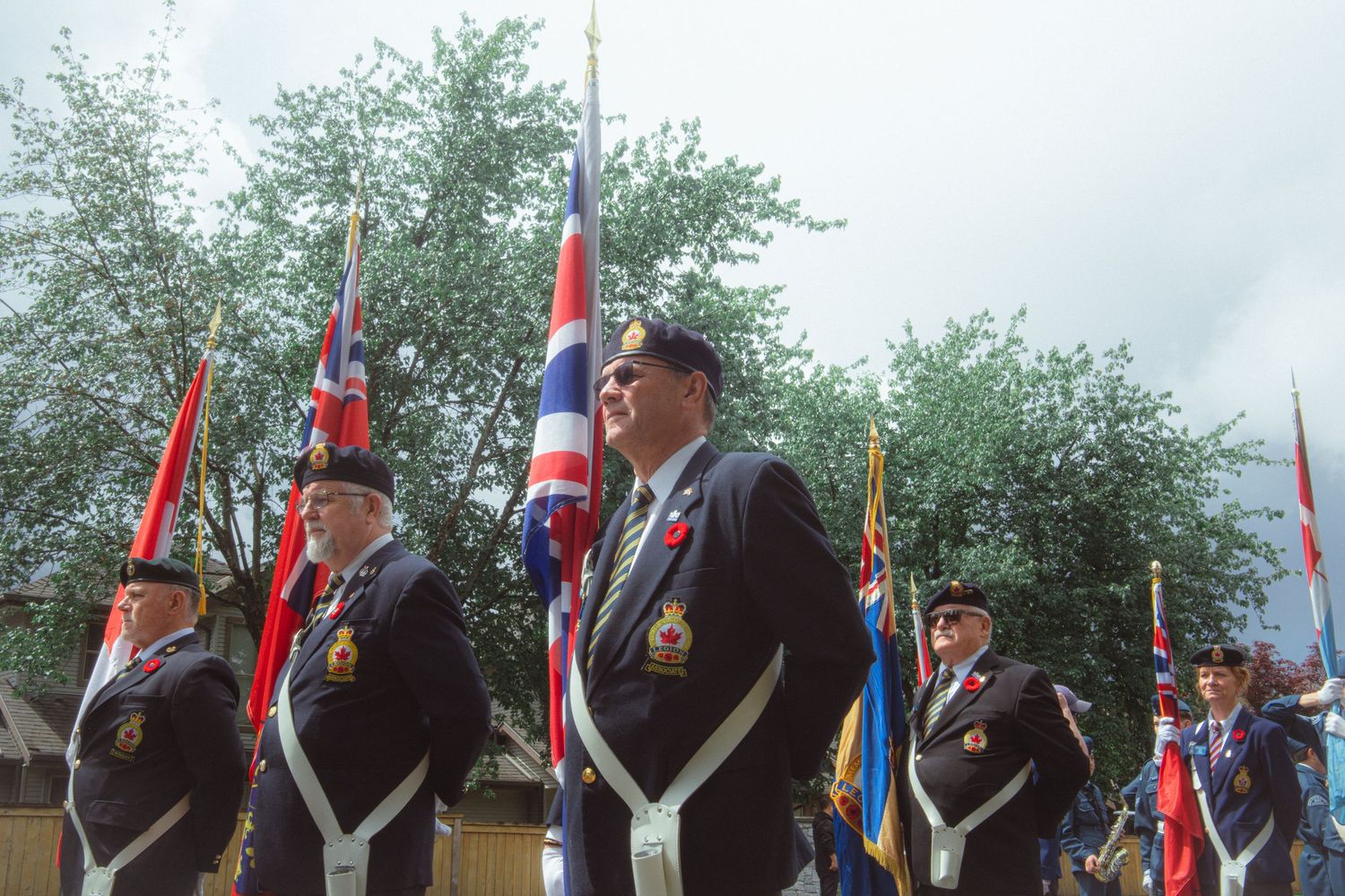 Veterans in uniform proudly carry flags including a Union Jack at an outdoor ceremony.