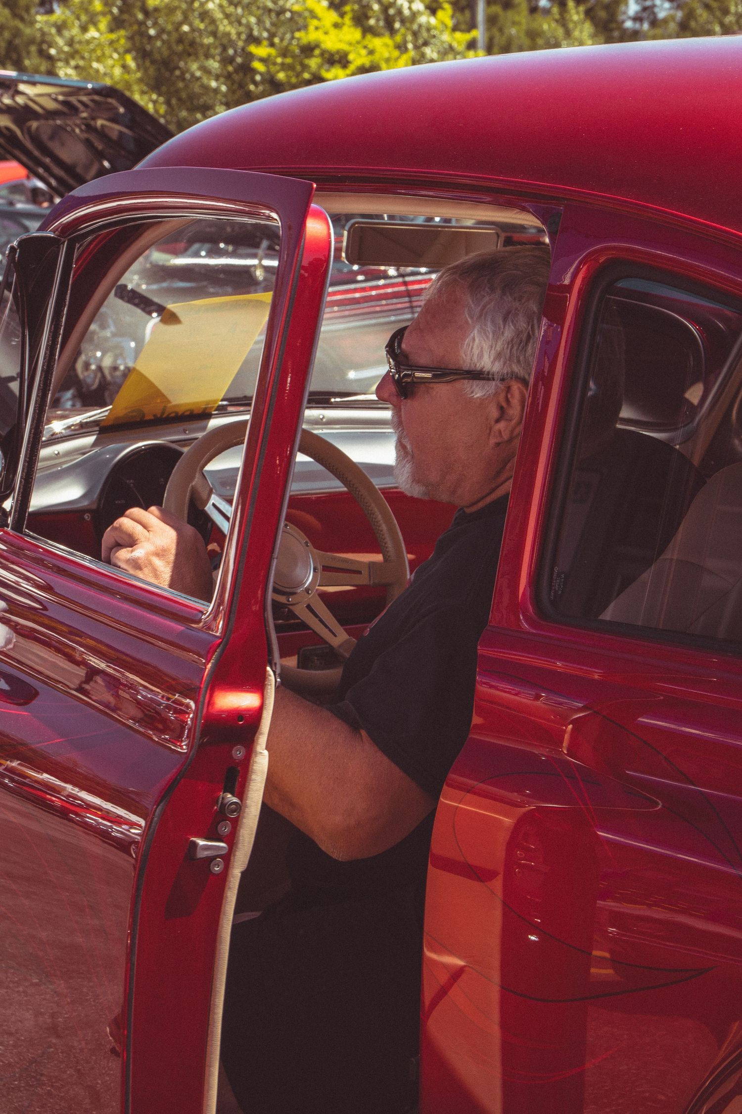 Senior man with sunglasses sitting behind the steering wheel of a restored classic red pickup truck at car show.