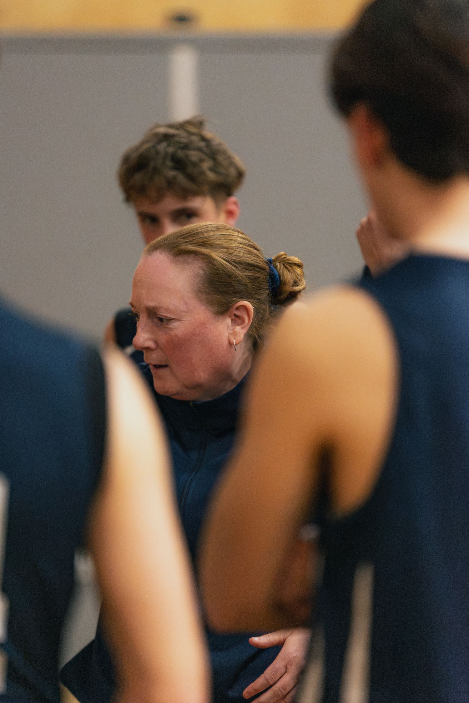 Female basketball coach in navy jacket gives focused instructions to players during a timeout huddle.