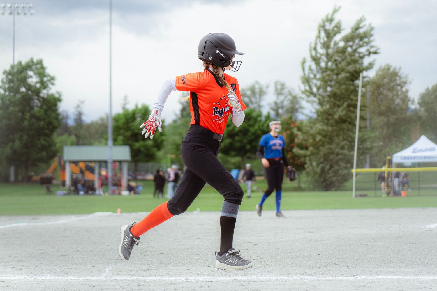 Youth softball player in orange uniform sprints toward first base during an outdoor game.