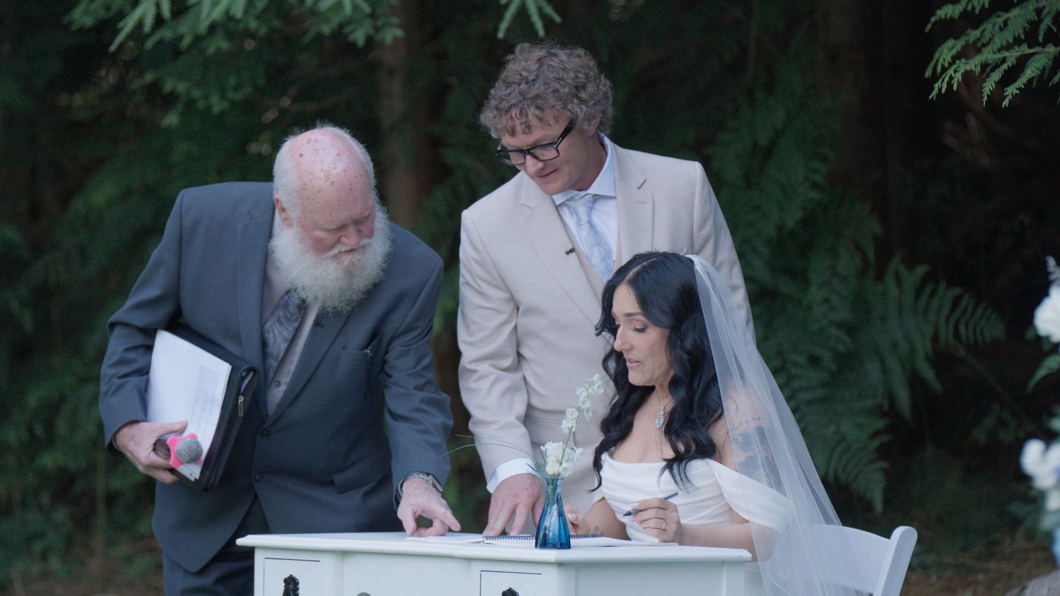 Bride and groom sign marriage certificate at white desk with officiant at outdoor wedding.