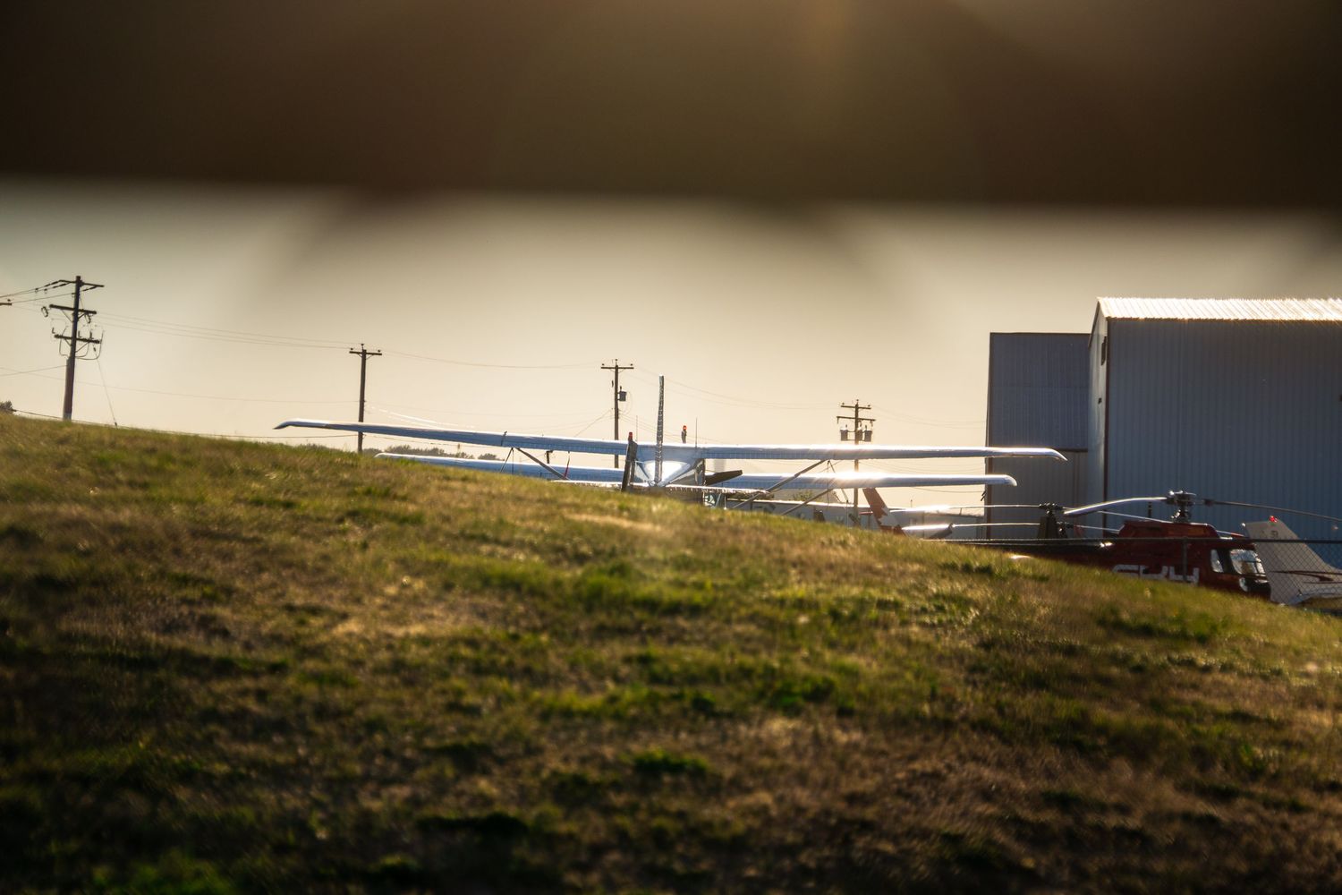A small aircraft parked on a grassy airfield at golden hour with utility poles and a building in the background.