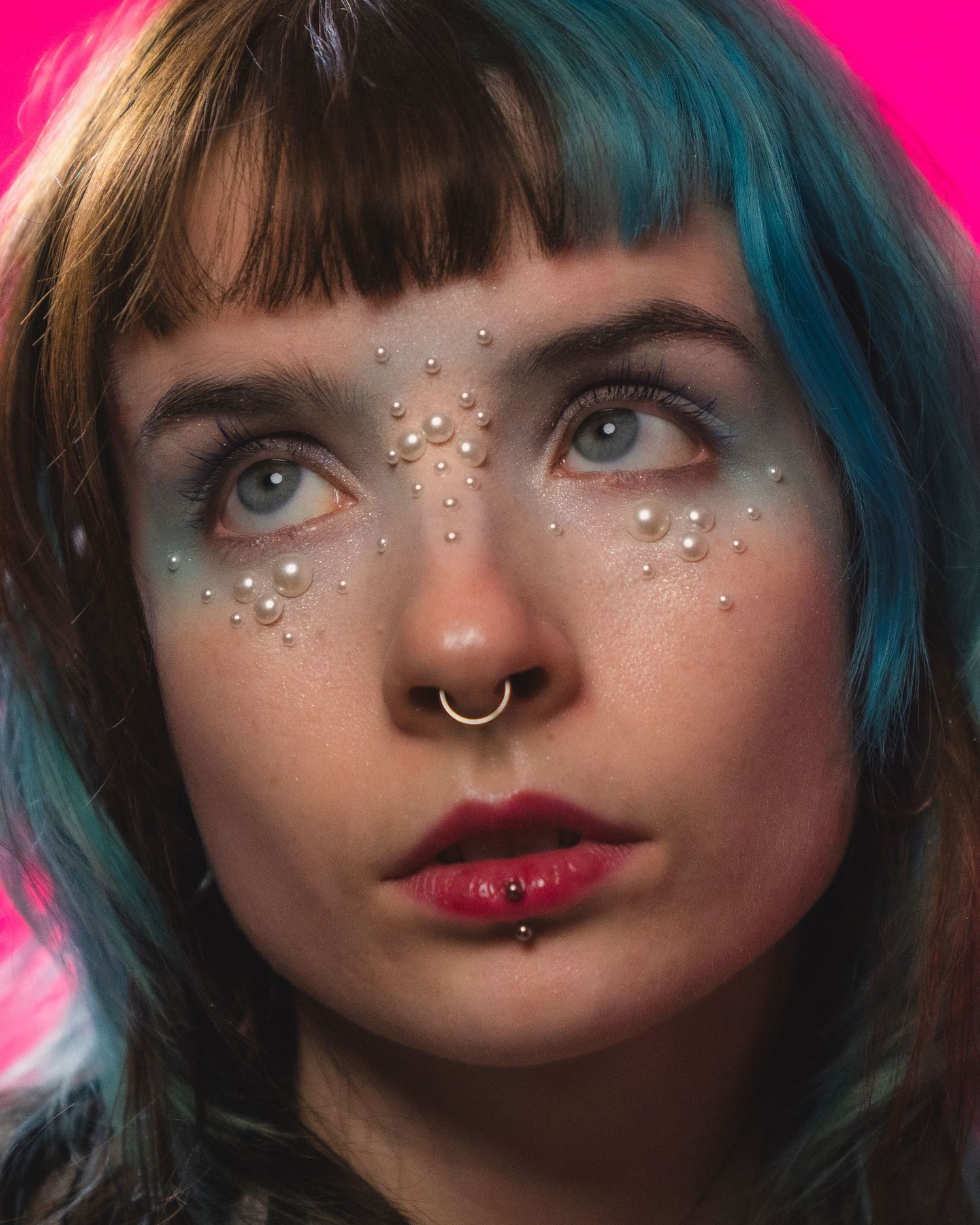 Close-up portrait of woman with teal hair, nose ring, and pearl face gems on pink background.