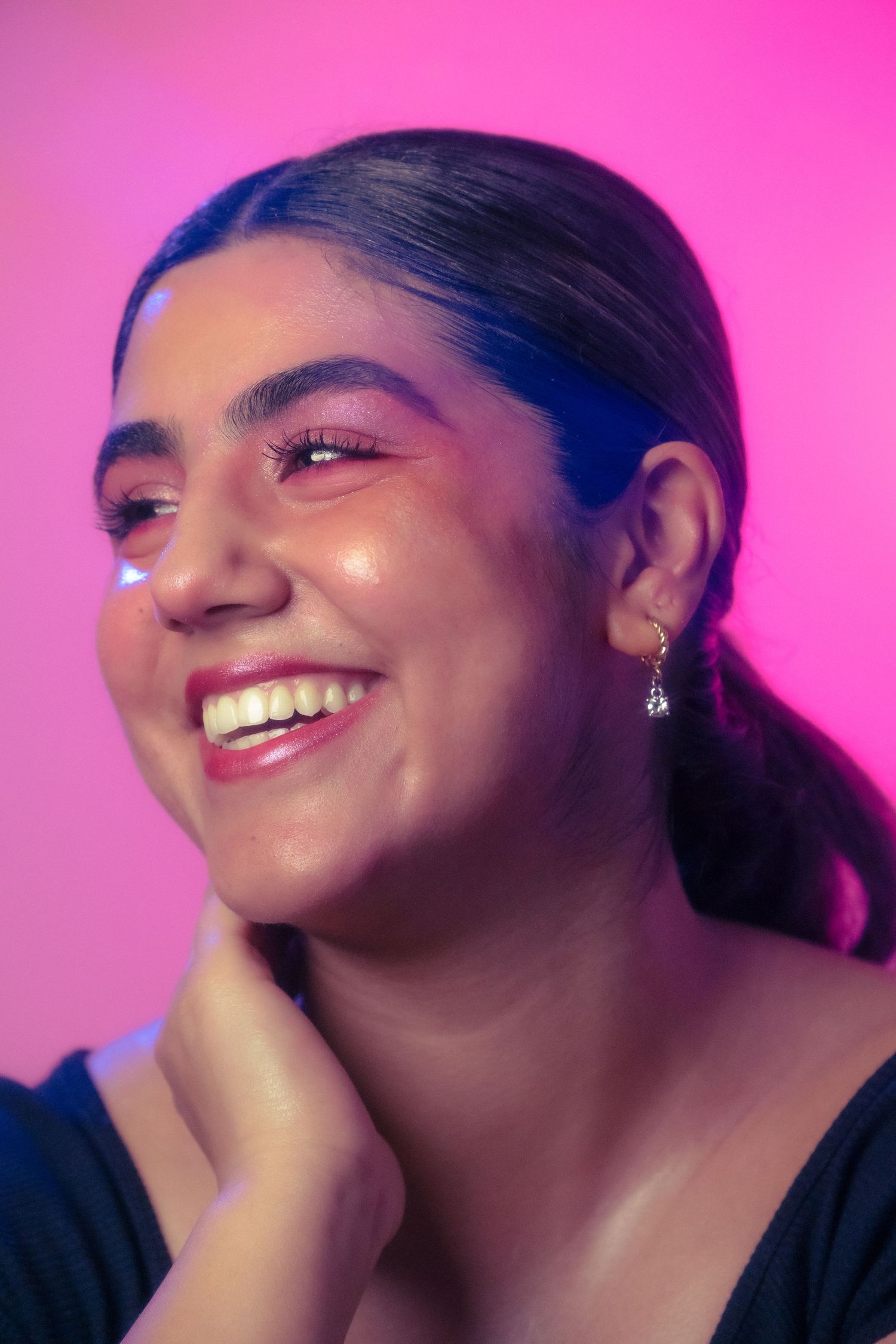 Laughing young woman with pink eyeshadow and red lips touching her neck against a vibrant pink neon backdrop.