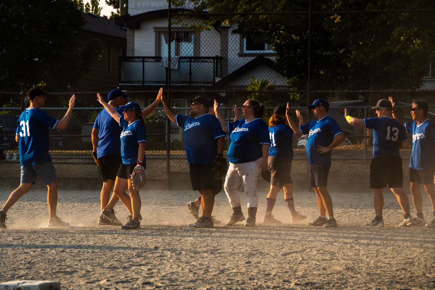 Softball team in blue uniforms celebrate with high-fives along the baseline after a game at sunset.