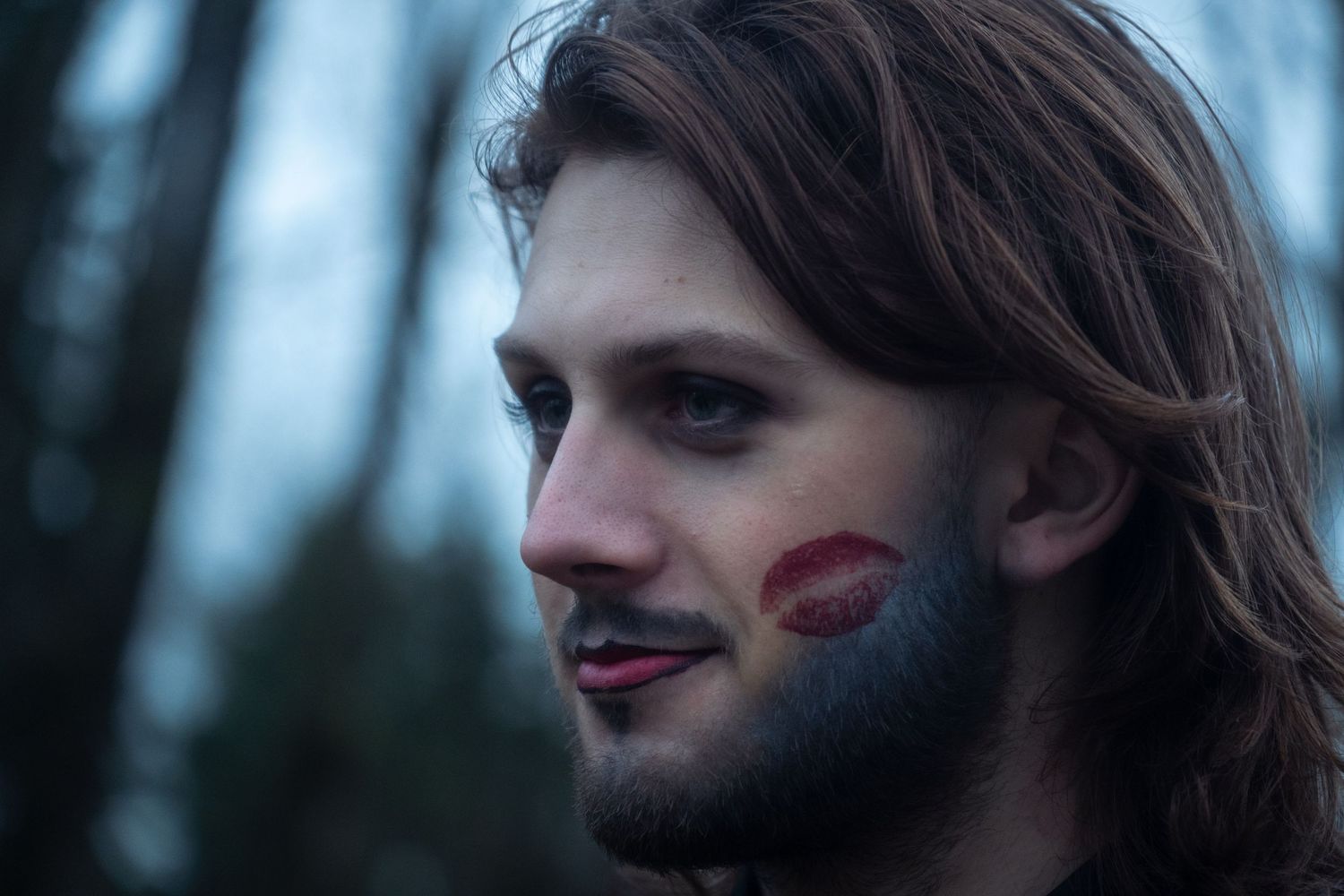Young man with long hair and red lipstick kiss mark on his cheek outdoors.