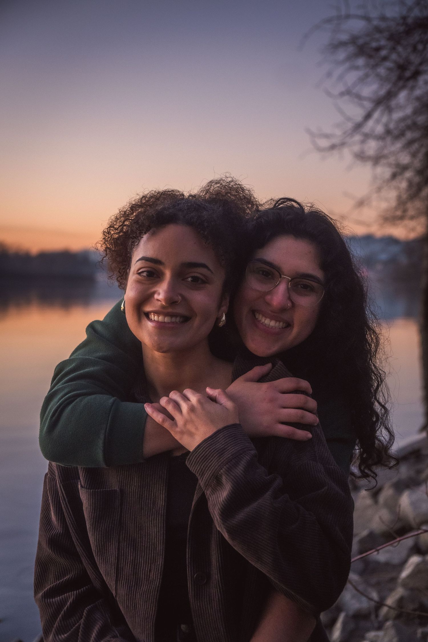 Two smiling women embrace warmly by a lakeside at dusk, sharing a joyful moment in the golden sunset light.