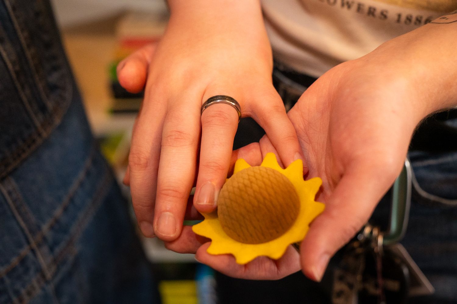 Two hands holding a small yellow sunflower-shaped object, one hand wearing a black ring.