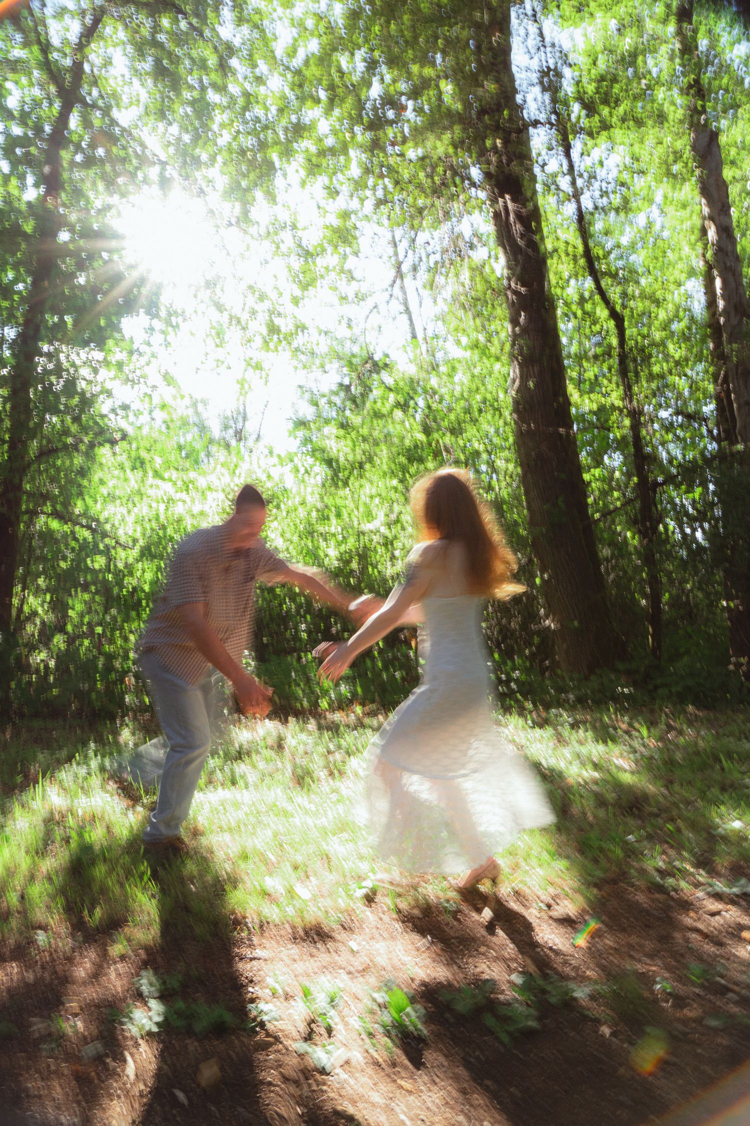 A couple dancing playfully in a sunlit forest, woman in white dress spinning with motion blur effect.