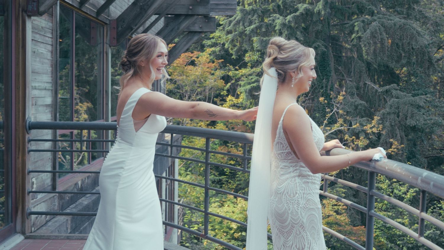 Two brides in white wedding dresses on a balcony surrounded by lush green trees.