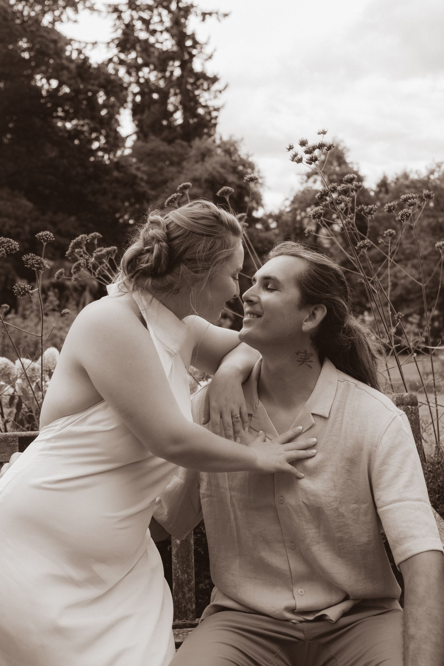 Sepia-toned photo of bride and groom sharing a tender moment on a bench surrounded by wildflowers.