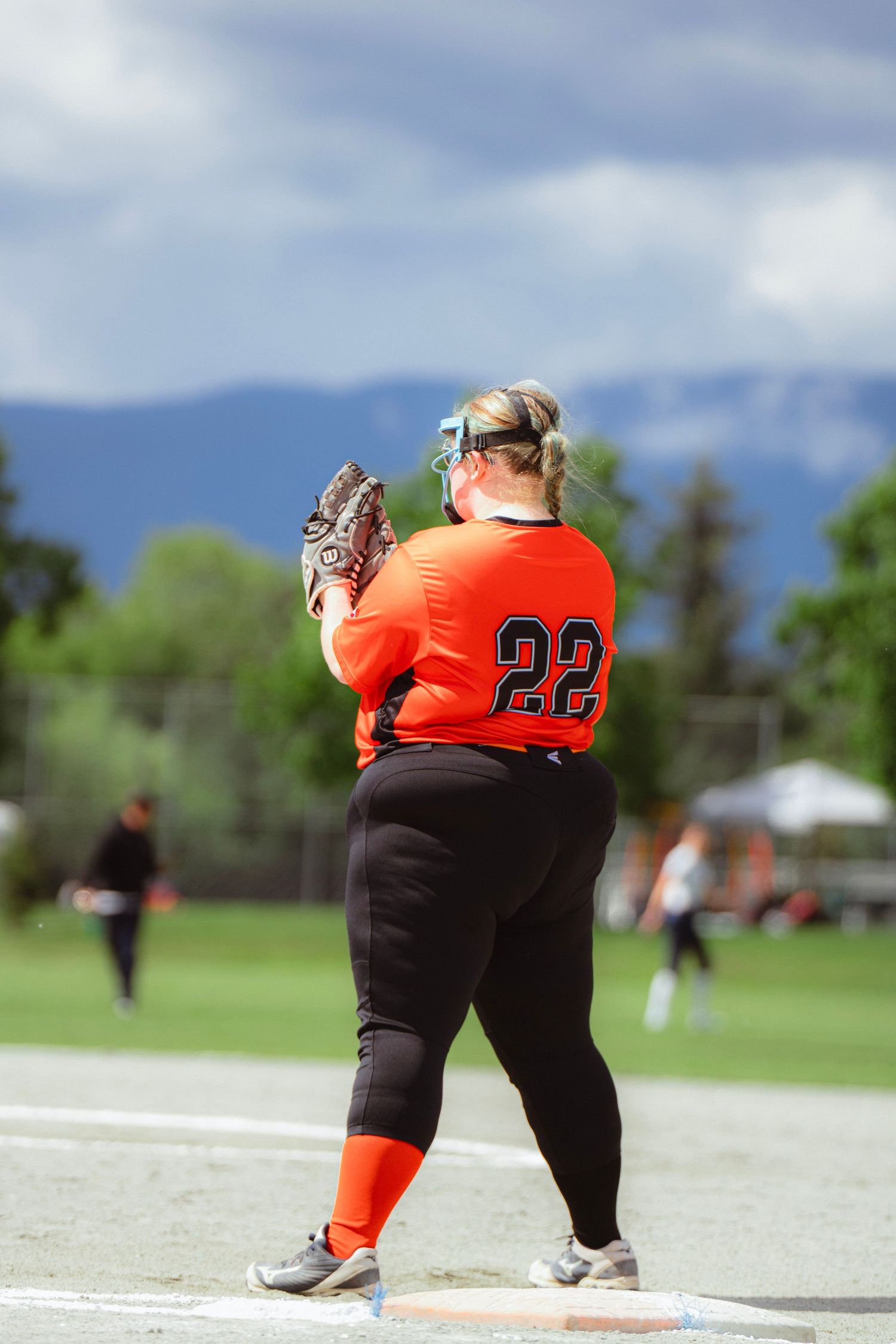 Softball player number 22 in orange jersey prepares to throw from first base with mountains behind.