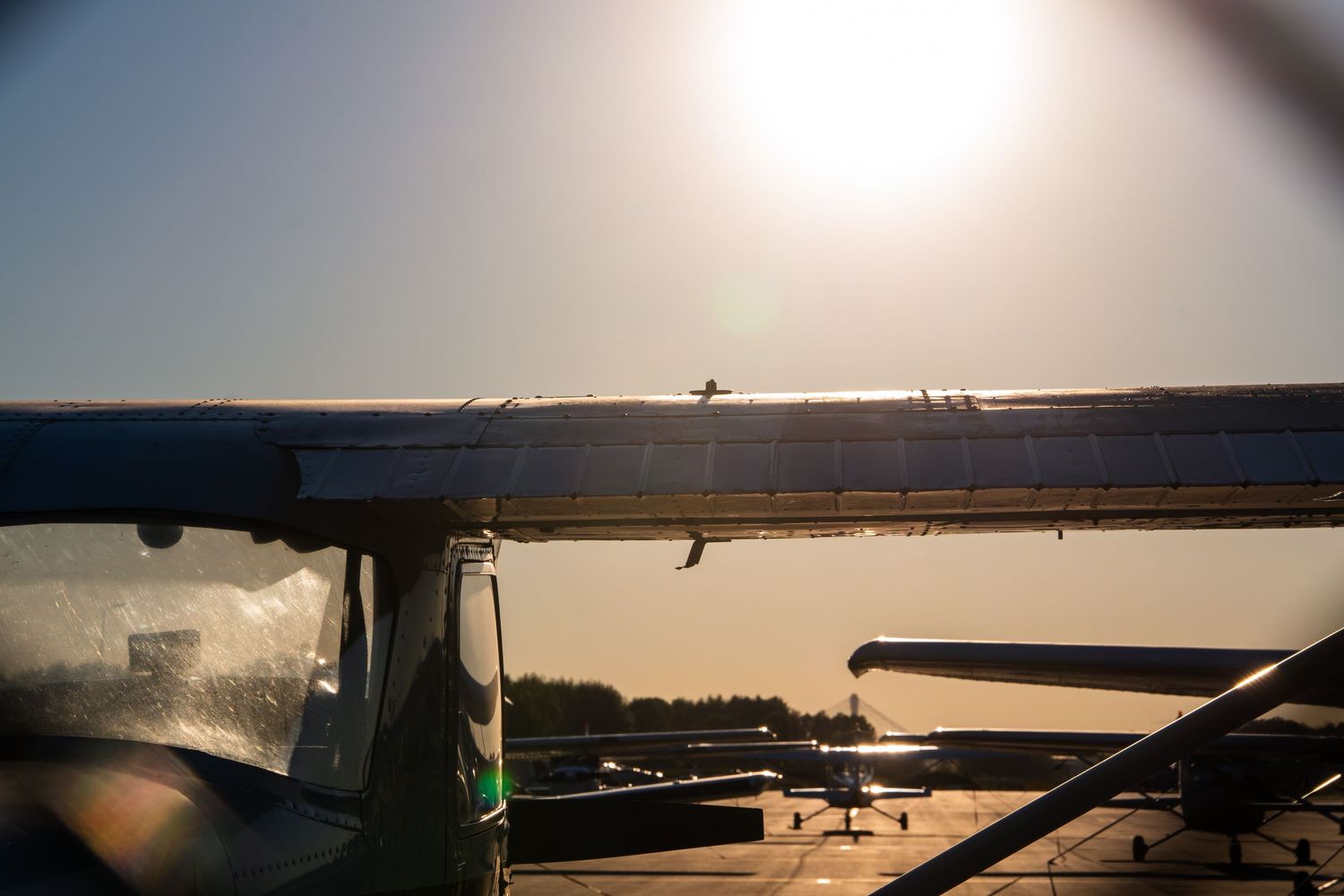 Close-up of a small aircraft wing at sunset with other planes and a calm waterway visible in the background.