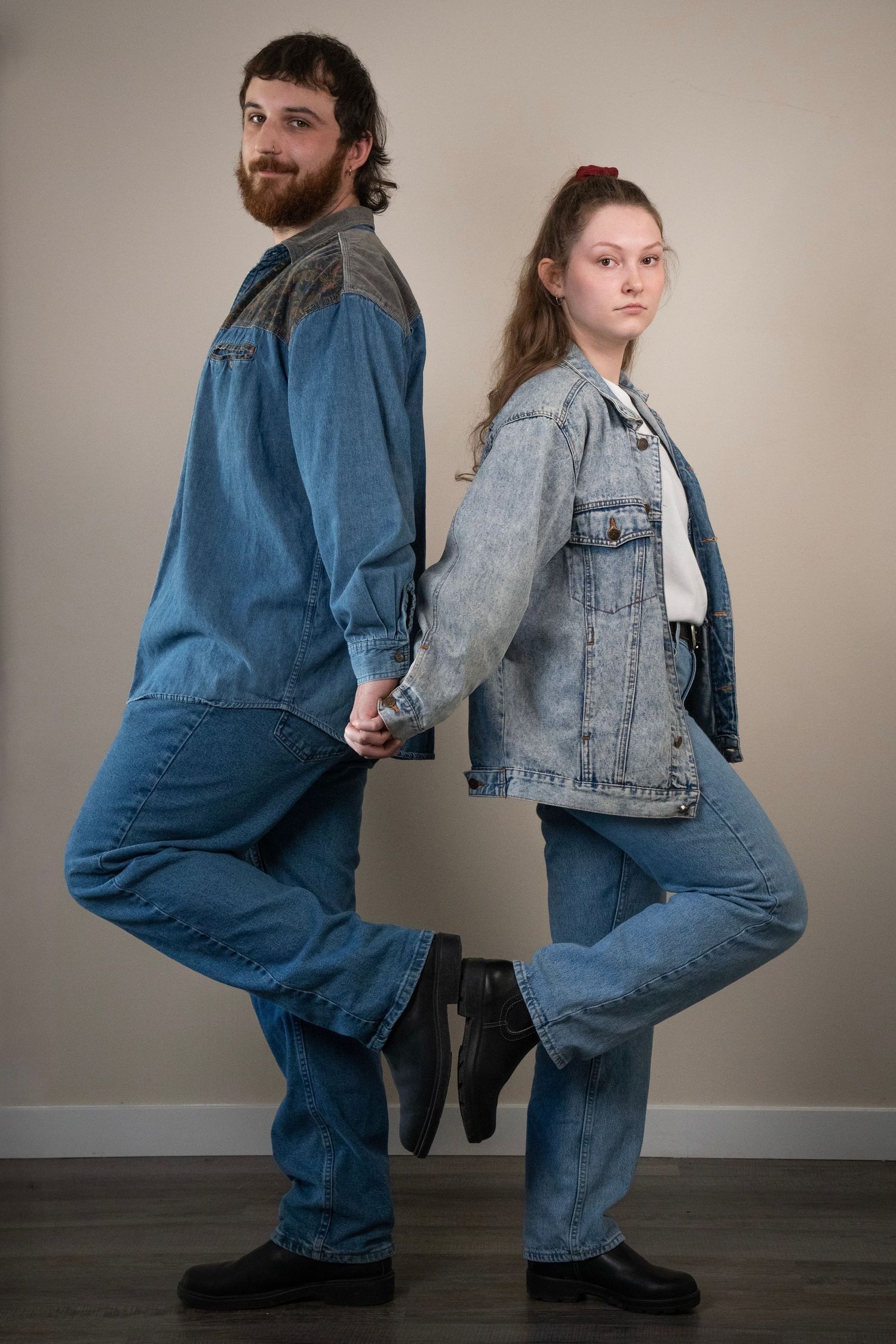 A stylish couple in coordinating denim outfits stand back-to-back holding hands against a neutral background.