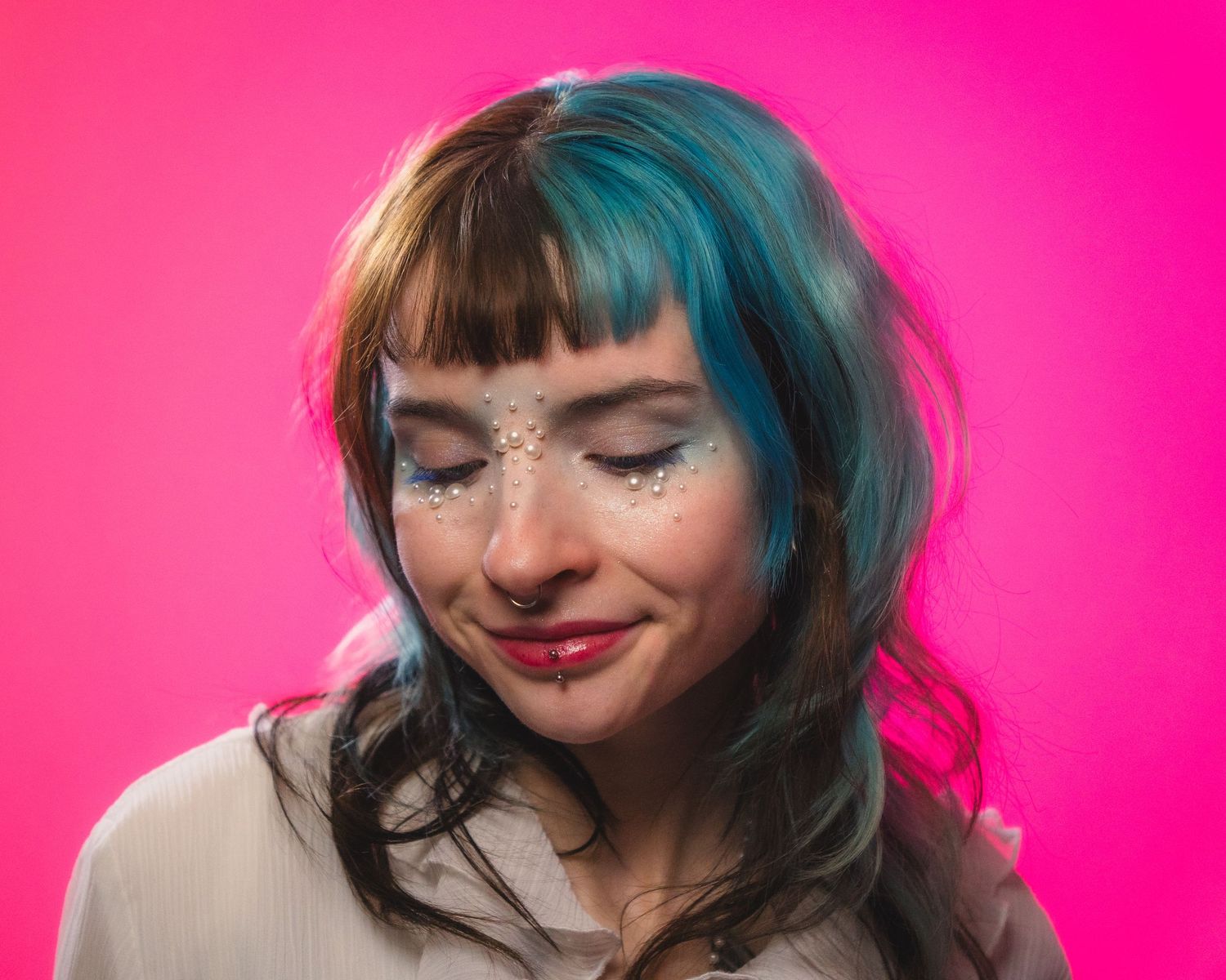Woman with teal hair and glitter face gems smiling with eyes closed on pink background.