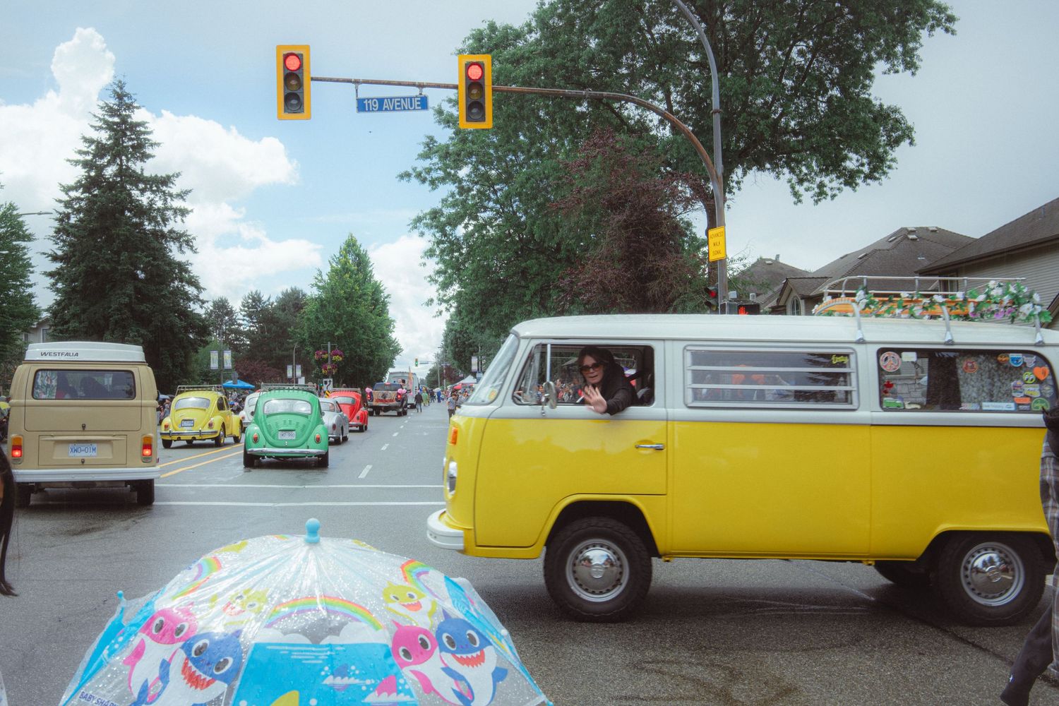 A yellow VW camper van leads colorful Volkswagen Beetles in a sunny street parade.