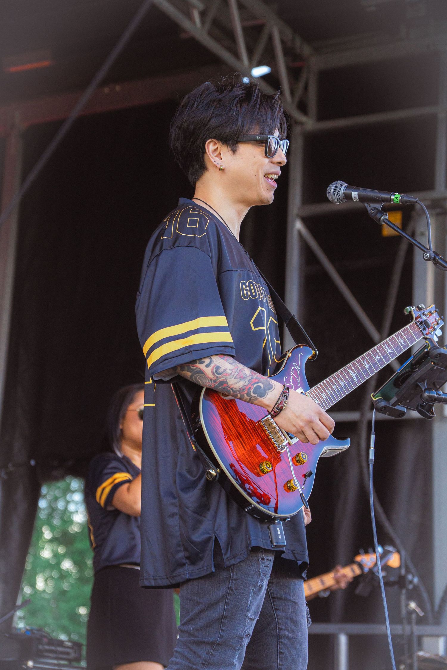 Tattooed guitarist in black jersey performing on outdoor stage with microphone at festival.