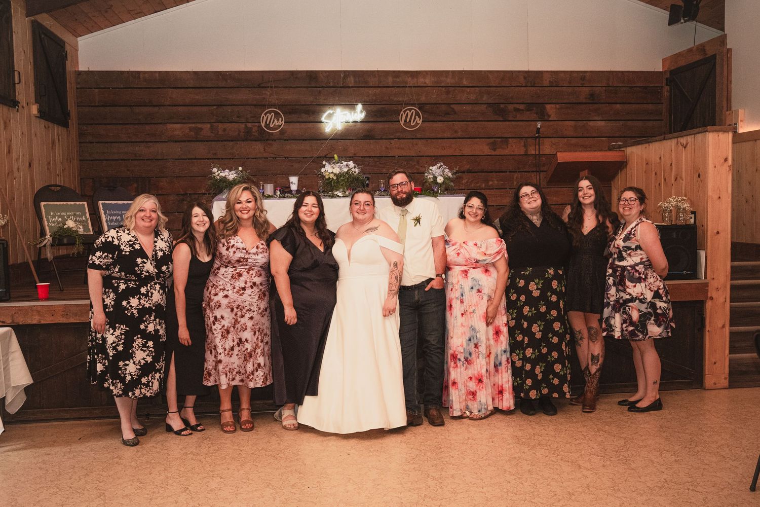 Bride and groom pose with a group of female guests in floral dresses at barn wedding.