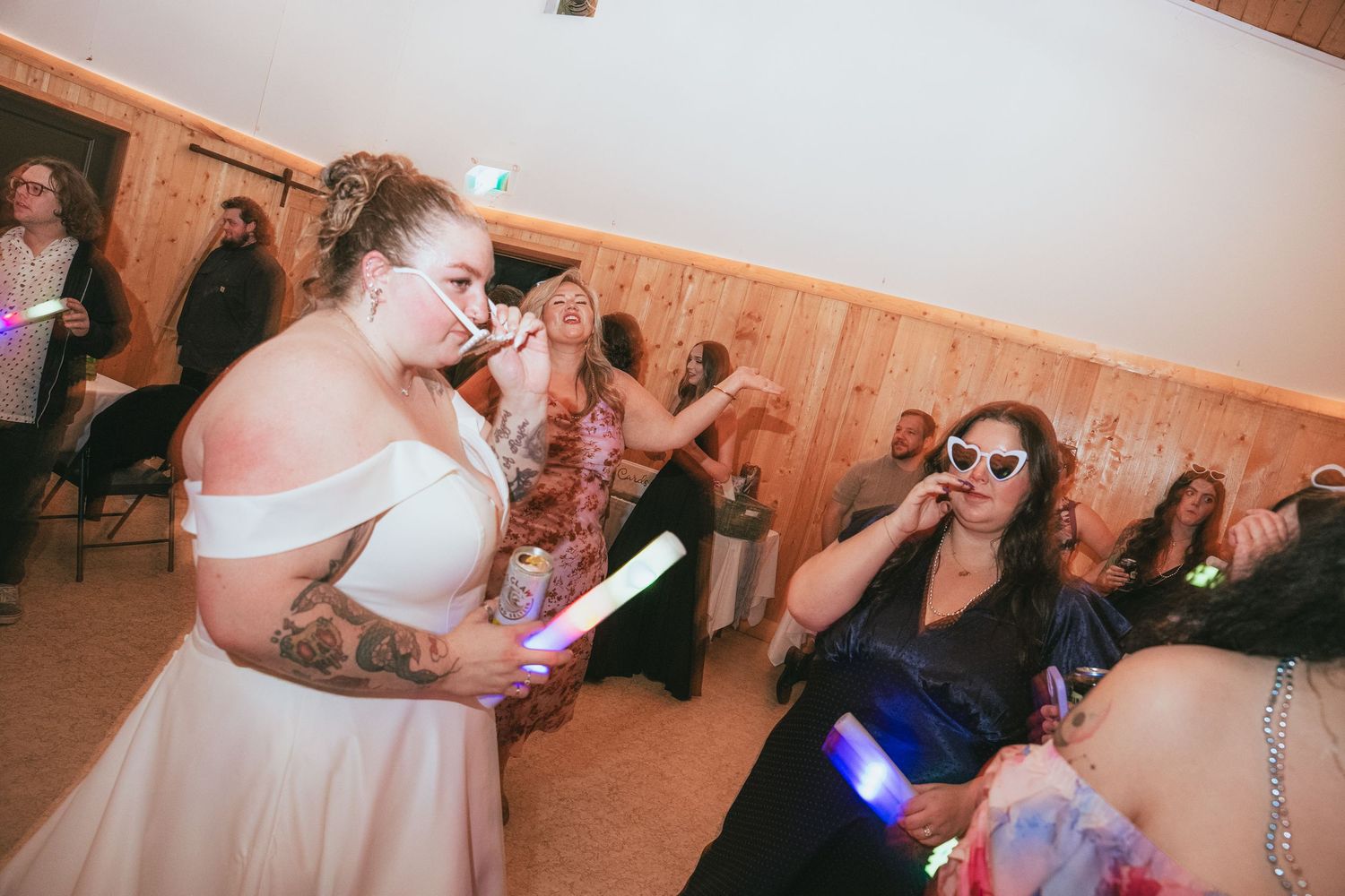 Bride in white off-shoulder dress with tattoos dances with guests holding glow sticks at a lively wedding reception.