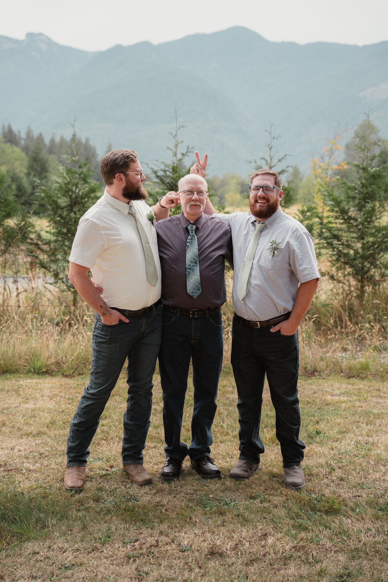 Three bearded men posing outdoors at a wedding with mountain scenery, two giving bunny ears to the man in the middle.