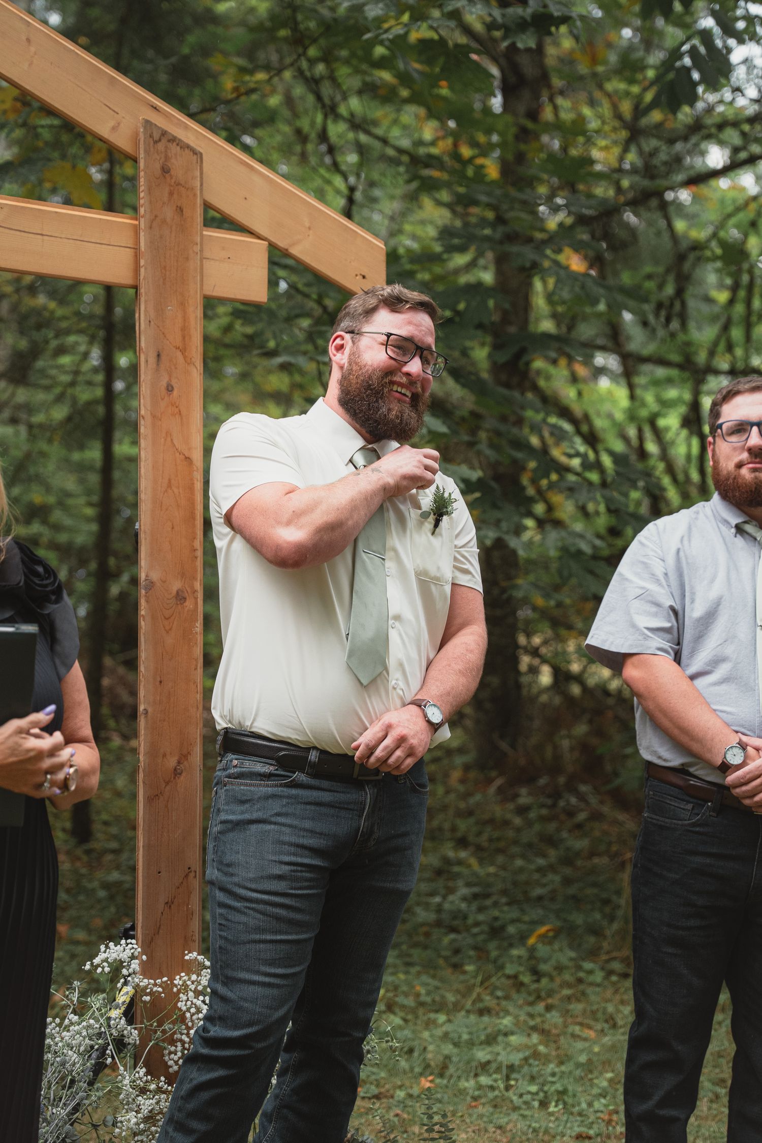 Bearded groom in white shirt and sage tie laughs joyfully at outdoor woodland wedding ceremony altar.