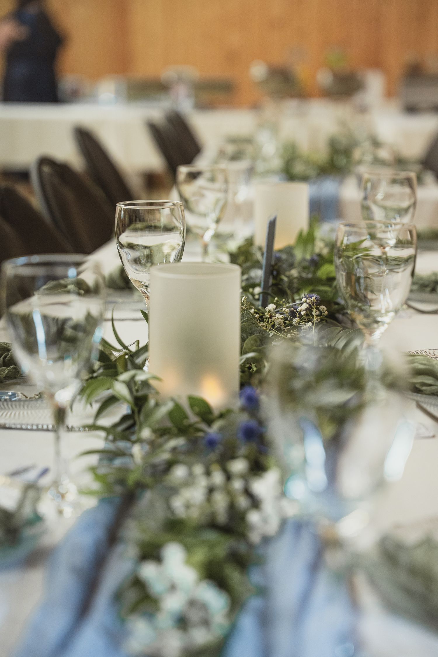 Elegant wedding table setting with greenery runner, white candles, and crystal glasses on a light blue tablecloth.