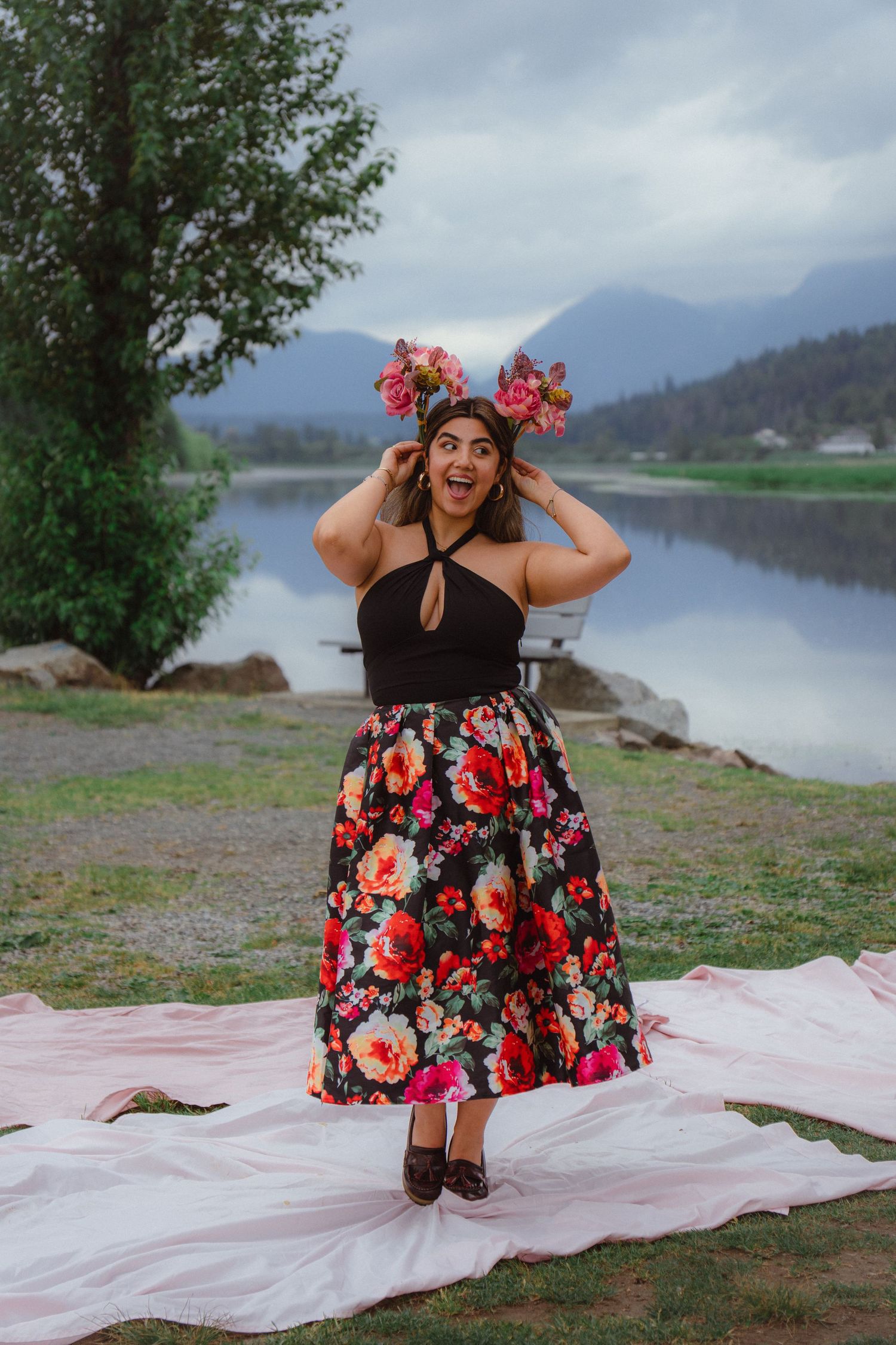 Woman in floral skirt placing flowers in hair laughing by a scenic mountain lake backdrop.