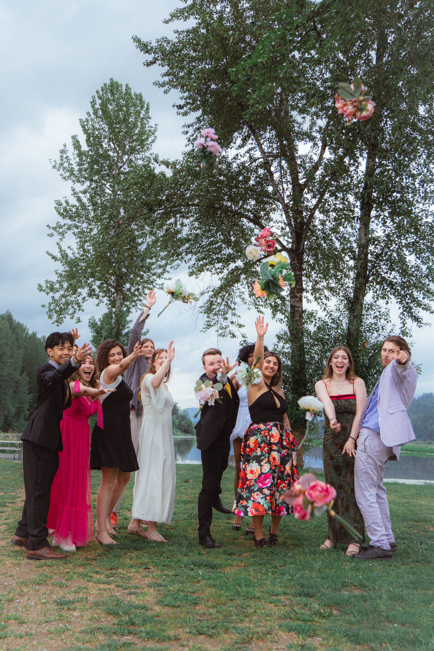 Wedding party joyfully tossing colorful bouquets in the air outdoors near trees.