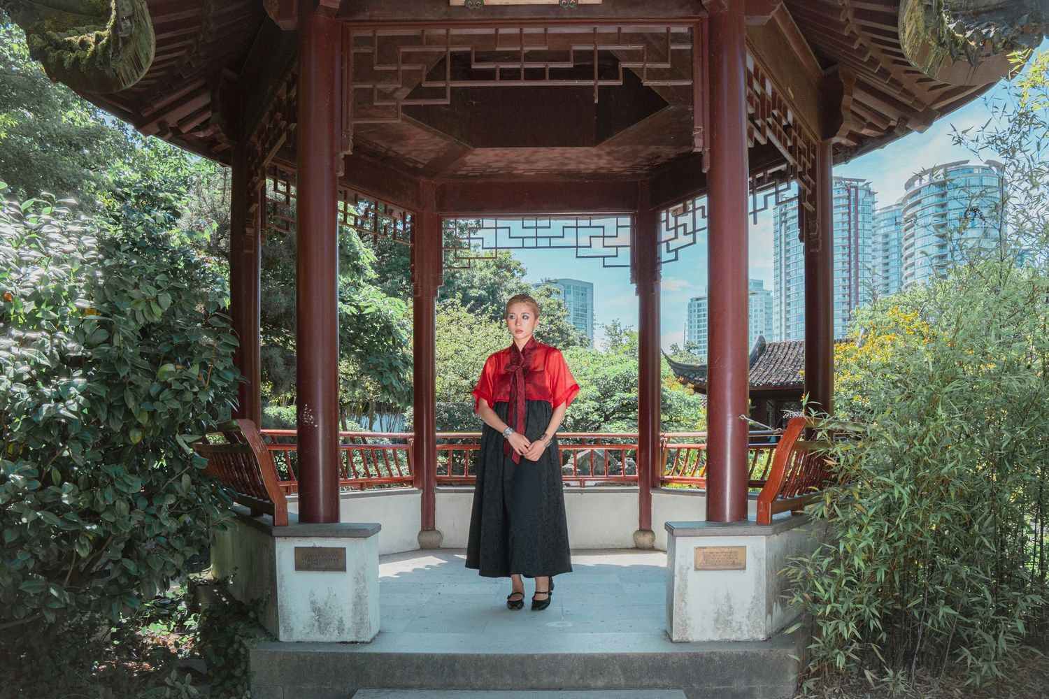 Woman in red top and black skirt stands in a traditional Chinese wooden pavilion surrounded by lush greenery.